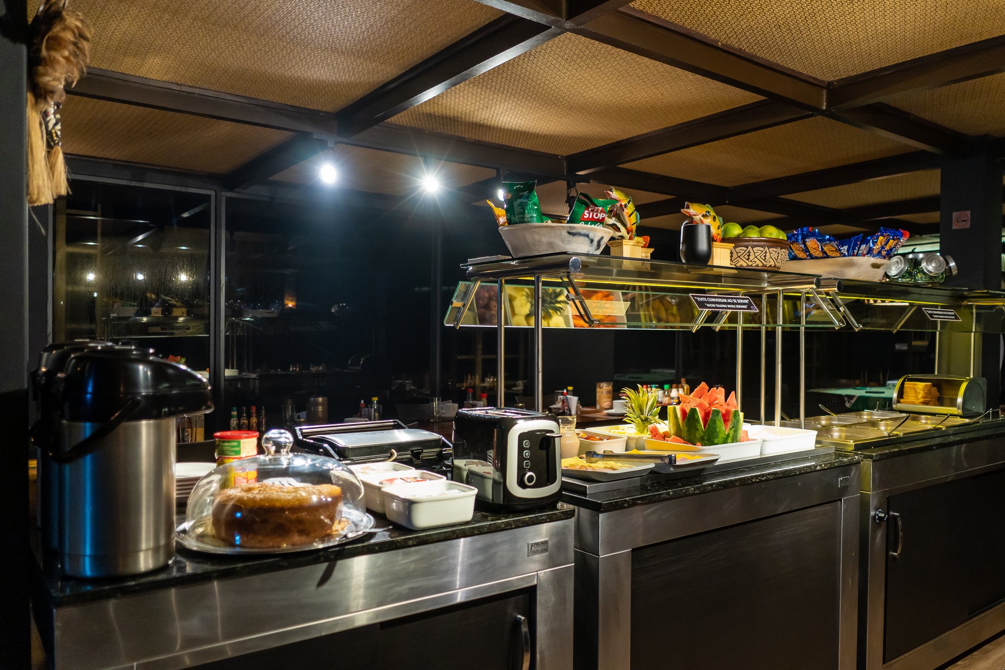 A well-lit onboard buffet on a luxury Amazon cruise in Brazil, featuring sliced watermelon, fresh tropical fruits, a whole cake under a glass dome, a toaster, thermal coffee urn, and snacks on tiered shelving, with dark river views through large windows at night
