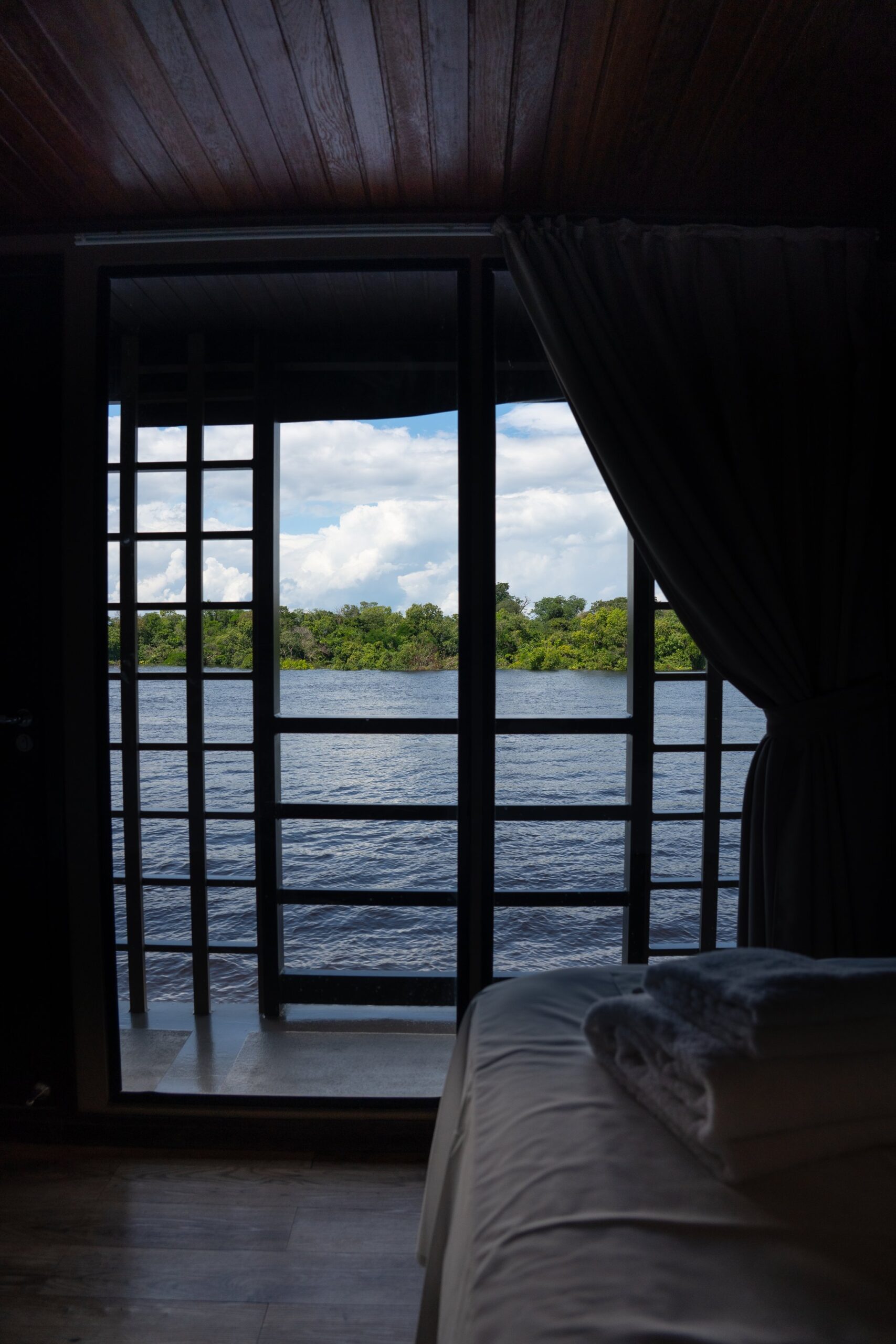 View from a dark, wood-panelled suite cabin aboard a luxury Amazon cruise in Brazil, with floor-to-ceiling grid windows framing a wide river and lush green treeline under a cloudy sky