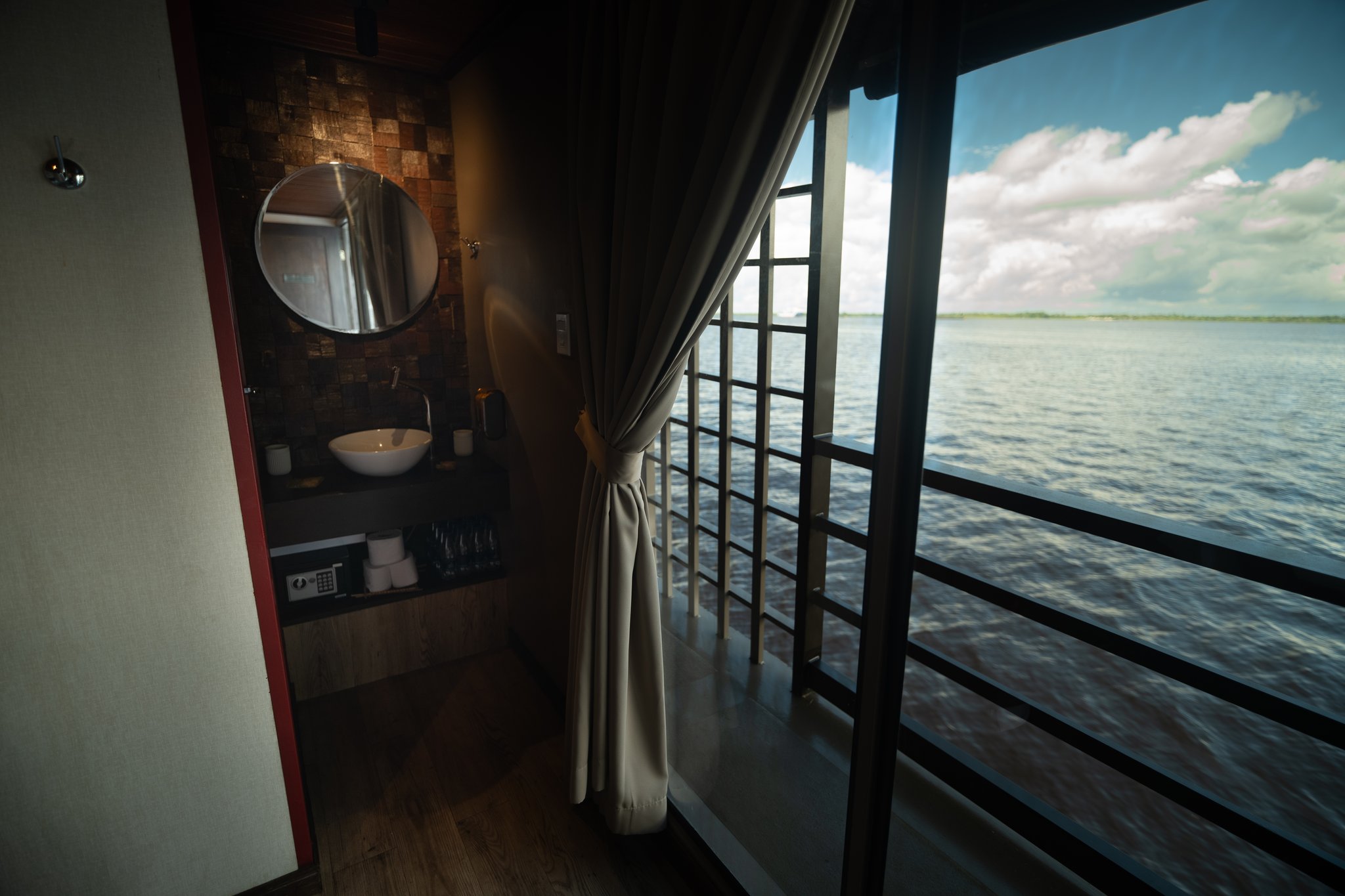 Suite cabin interior on a luxury Amazon cruise in Brazil featuring a round mirror over a vessel-sink vanity, room safe, and floor-to-ceiling windows opening onto the wide river under a blue sky