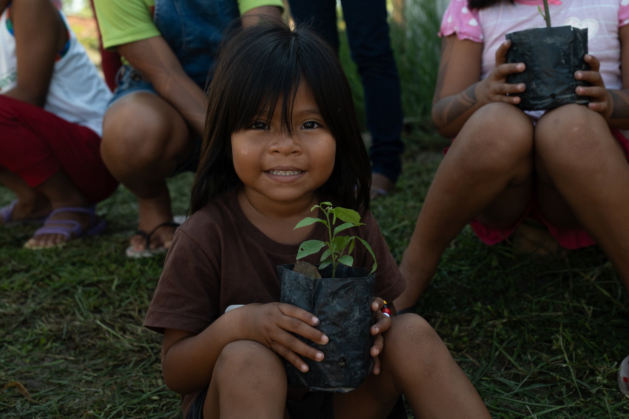 A smiling young girl holds a green seedling in a black nursery pot during a community visit on a luxury Amazon cruise in Brazil, with other children seated on the grass behind her