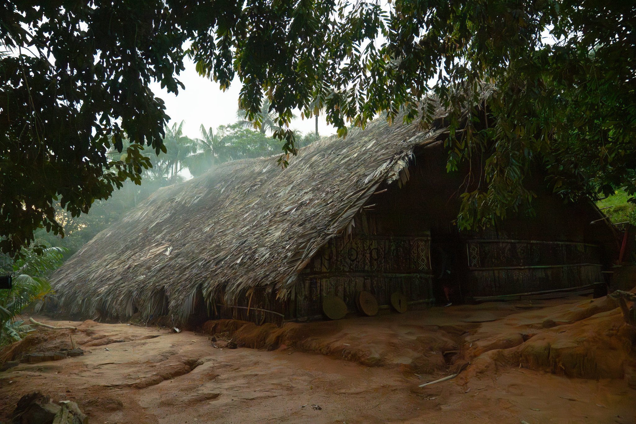 A large palm-thatched indigenous communal longhouse with geometric painted decorations on its wooden walls, surrounded by tropical trees and red-earth ground on a Luxury Amazon Cruise in Brazil