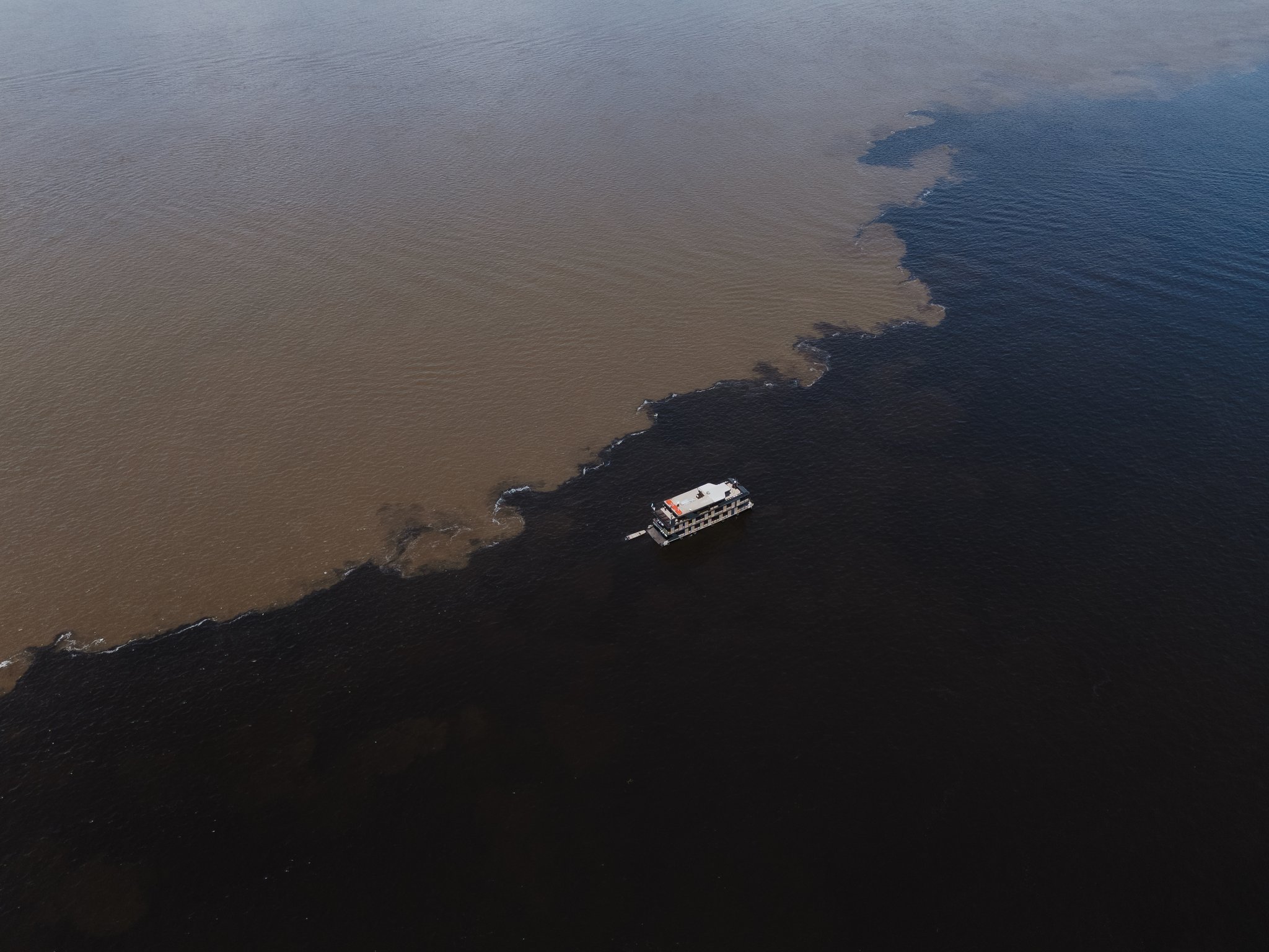 Aerial view of a riverboat crossing the Meeting of Waters on a luxury Amazon cruise in Brazil, where dark black-water and sandy brown-water rivers meet in a sharp, winding boundary across the frame