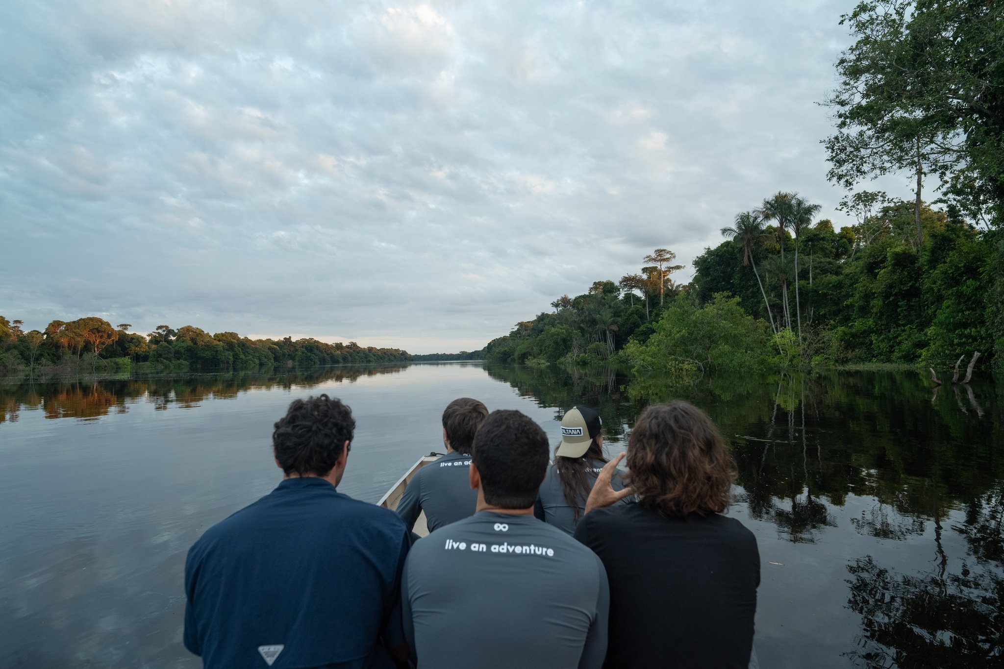 Four travellers seated at the bow of a small boat on a calm, dark-water river, surrounded by dense rainforest canopy on both sides during an Amazon river cruise in Brazil