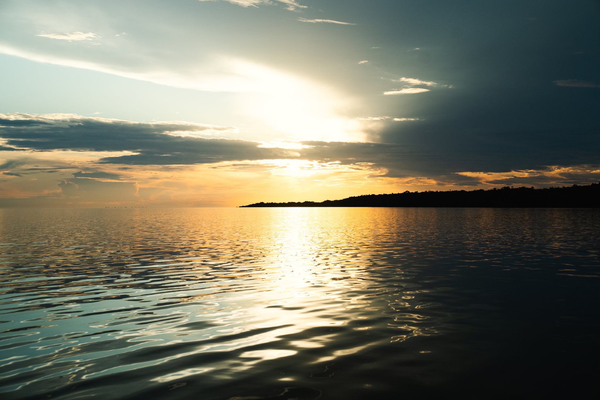 Golden sunset reflected across a vast, calm river surface on a luxury Amazon cruise in Brazil, with a dark forested island silhouetted on the horizon beneath dramatic clouds