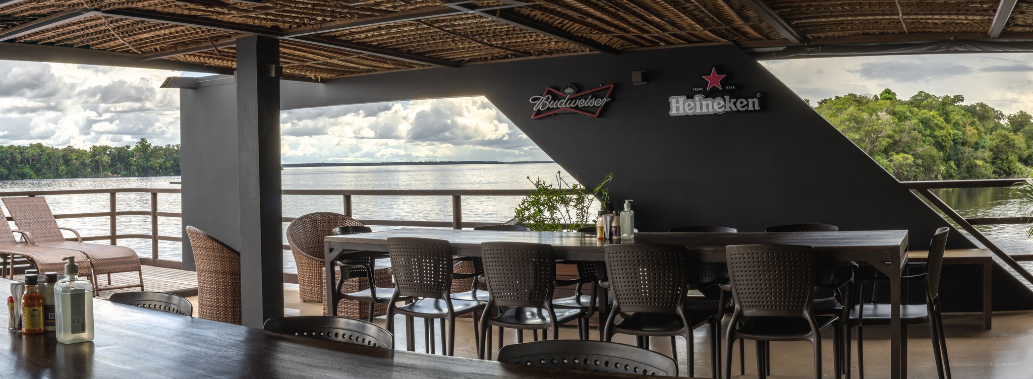 Open-air bar deck on a luxury Amazon cruise in Brazil, with rattan chairs, long dining tables under a woven-palm ceiling, and a wide river framed by dense jungle on both sides