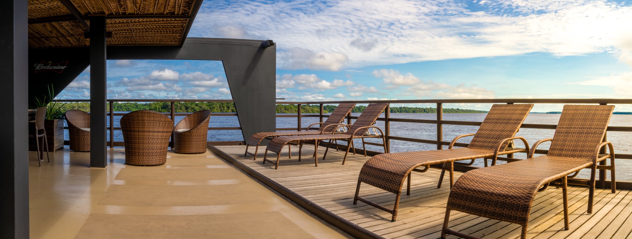 Wicker sun loungers and rattan chairs arranged on the upper sun deck of a luxury Amazon cruise in Brazil, overlooking a wide dark-water river lined with dense rainforest under a blue sky