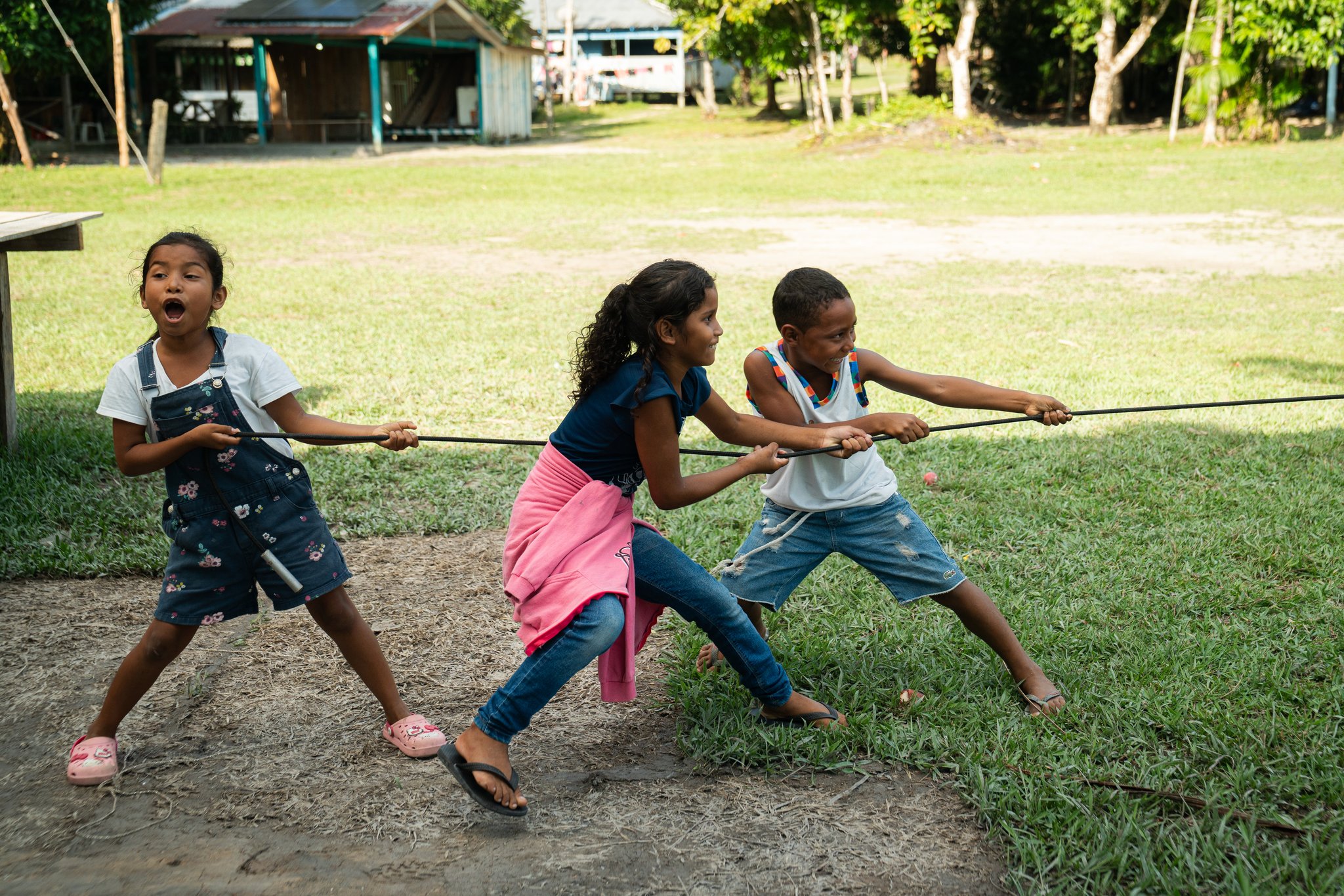 Three children laughing and straining on a rope in a tug-of-war game in a riverside village clearing, a highlight of community visits on a Luxury Amazon Cruise in Brazil
