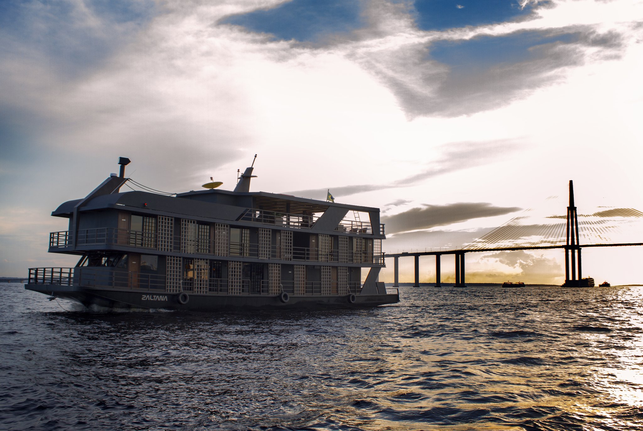 The Zaltana riverboat setting out on a luxury Amazon cruise in Brazil, with a cable-stayed bridge silhouetted against a dramatic sunset sky on the broad Rio Negro