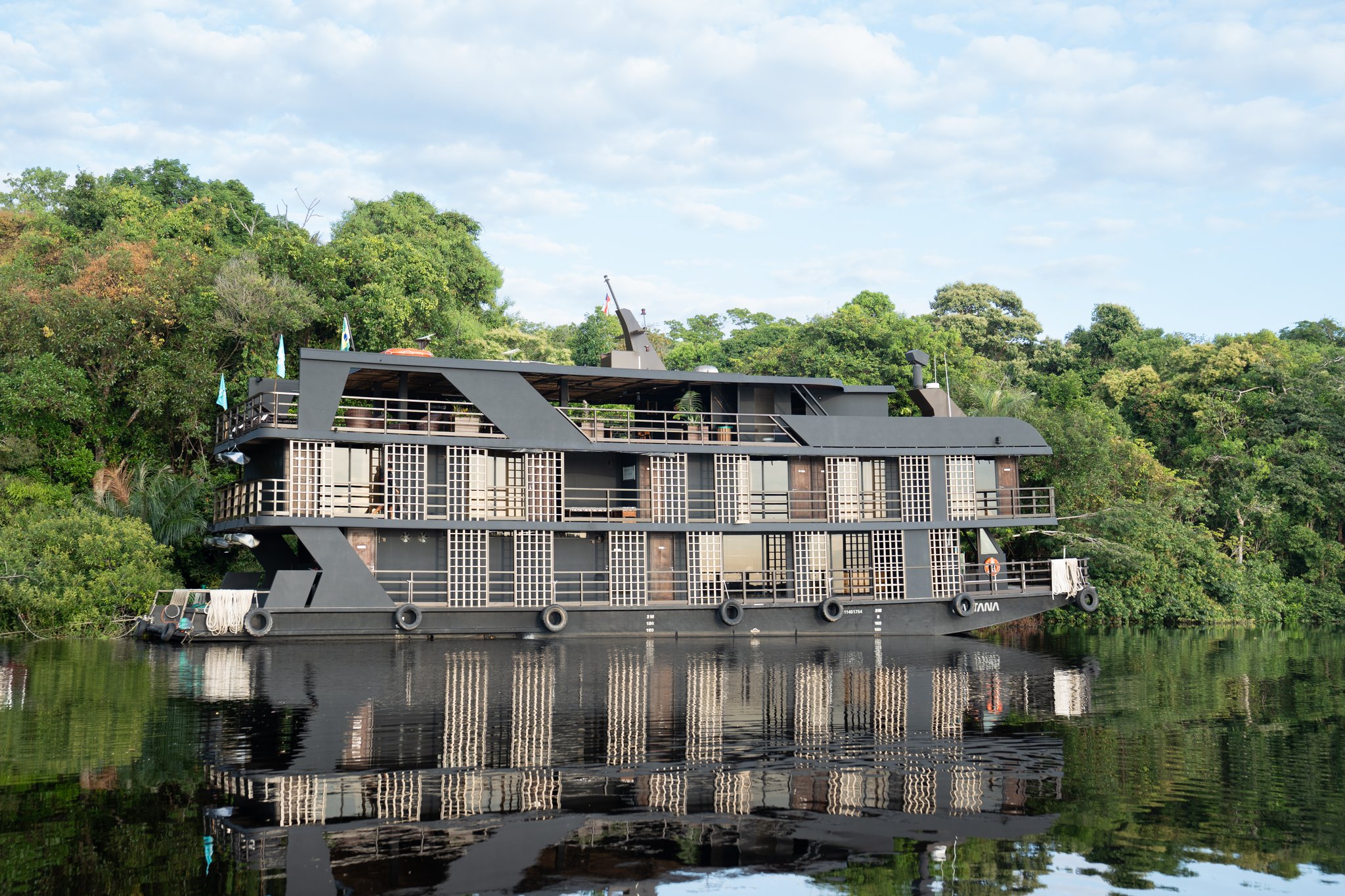 The Zaltana luxury Amazon cruise riverboat moored on a glassy dark-water river, its modern multi-deck dark hull and latticed cabin balconies reflected perfectly in the still water, backed by dense rainforest canopy under a partly cloudy sky