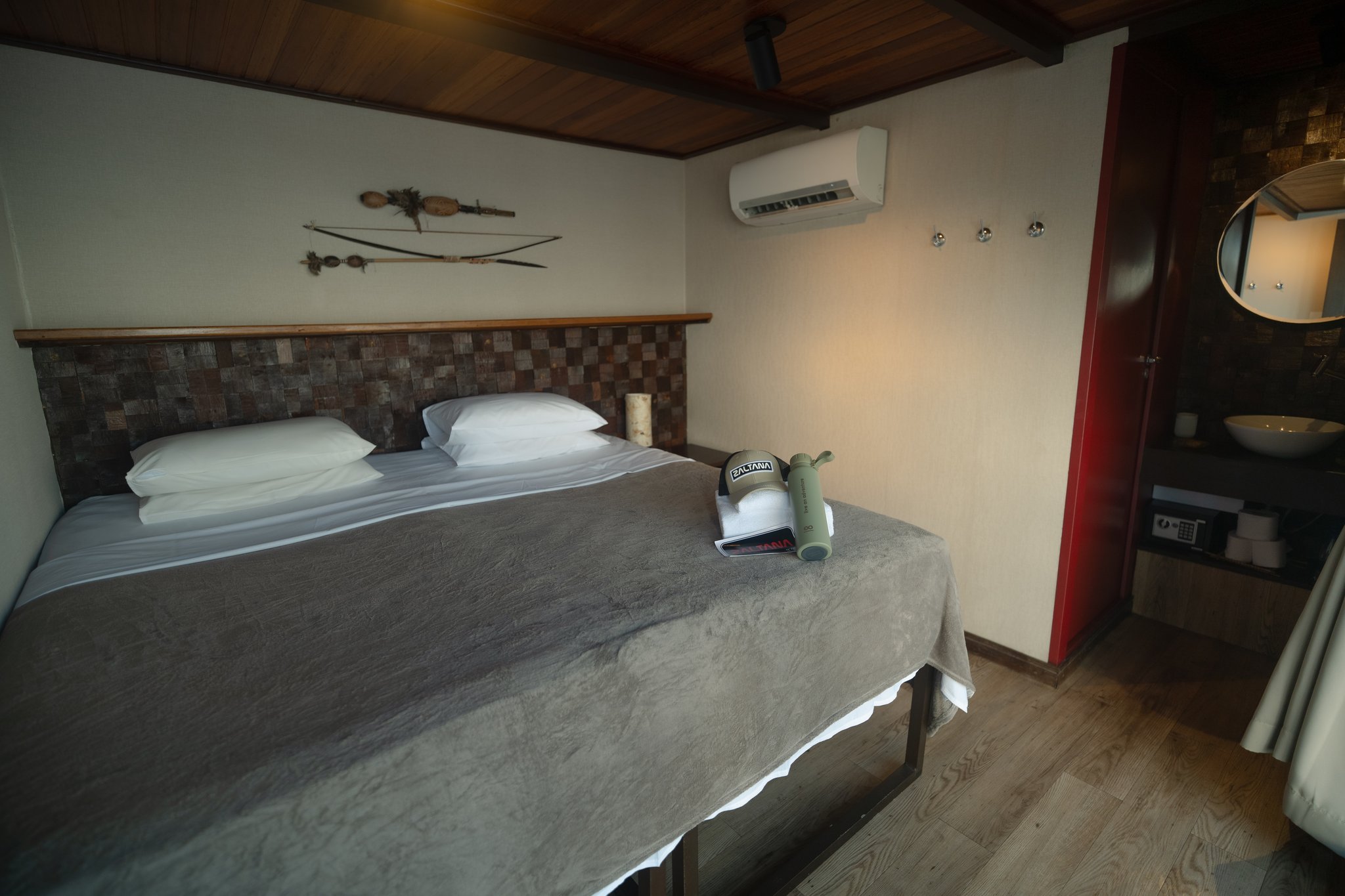 A neatly made double bed with white pillows and grey blanket inside a Luxury Amazon Cruise in Brazil suite cabin, featuring a mosaic wood headboard, indigenous bow-and-arrow wall art, wooden ceiling, air conditioning unit, and a binoculars case resting on the bed