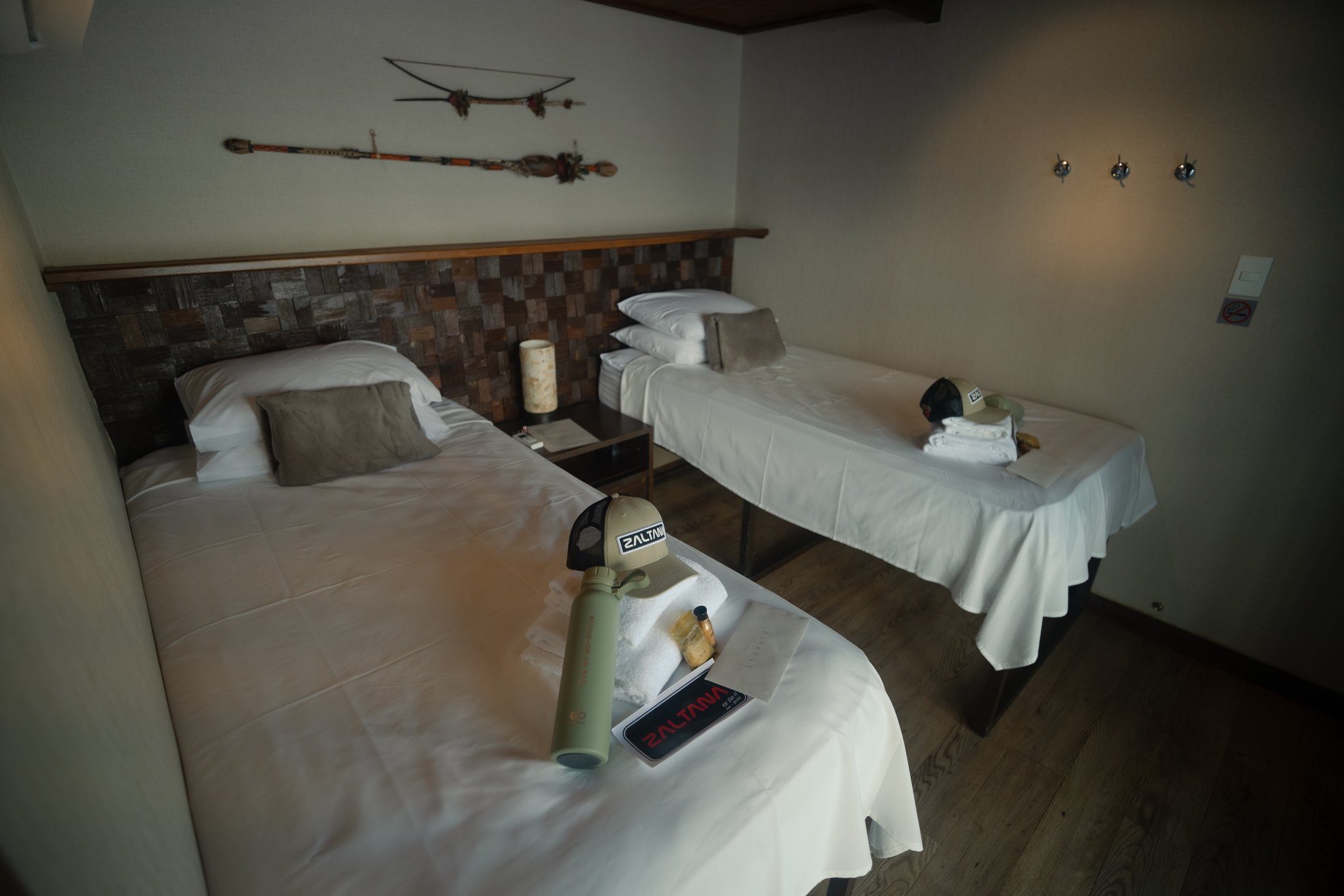 Twin-bed suite cabin aboard the Zaltana riverboat on a Luxury Amazon Cruise in Brazil, featuring crisp white linen, a stone-tile headboard, indigenous decorative artefacts on the wall, and Zaltana-branded guest amenities on the beds