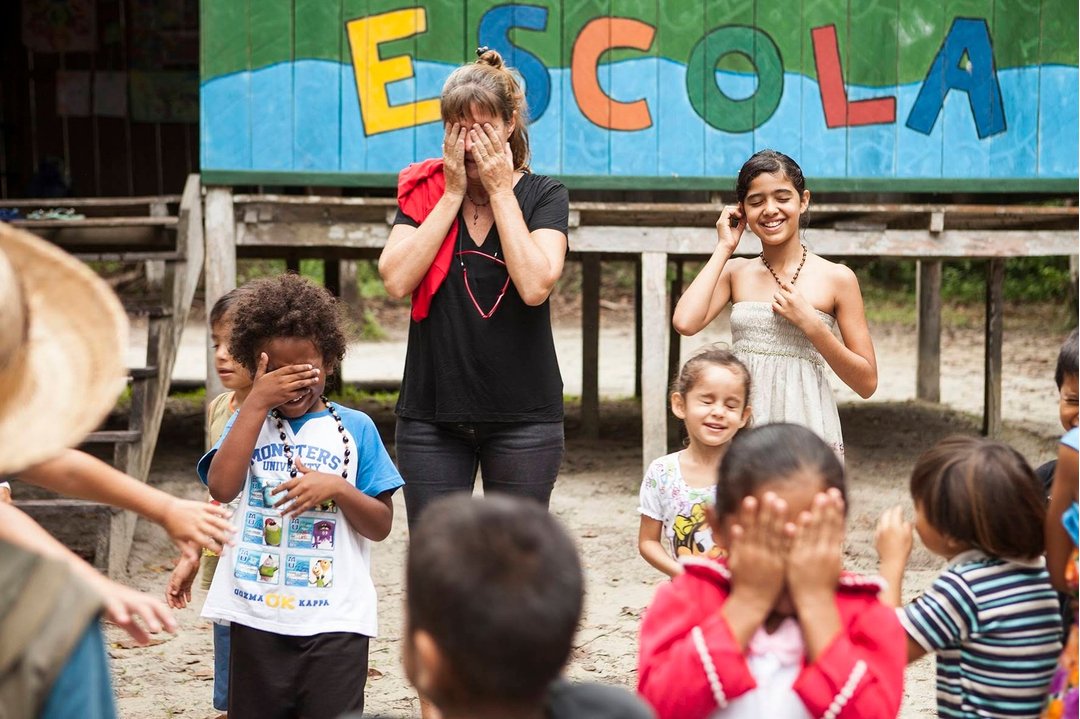 Laughter, covered eyes, and a game that needs no translation. A traveller and local children play a hand-covering game in front of a colourfully painted school building marked 'ESCOLA' during a community visit on a luxury Amazon lodge Brazil itinerary