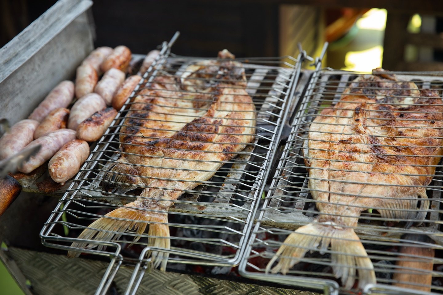 Charcoal, river fish, and sausages — lunch earned by the morning's adventure. Whole fish and sausages grilling in wire cages over charcoal at a luxury Amazon lodge Brazil outdoor barbecue