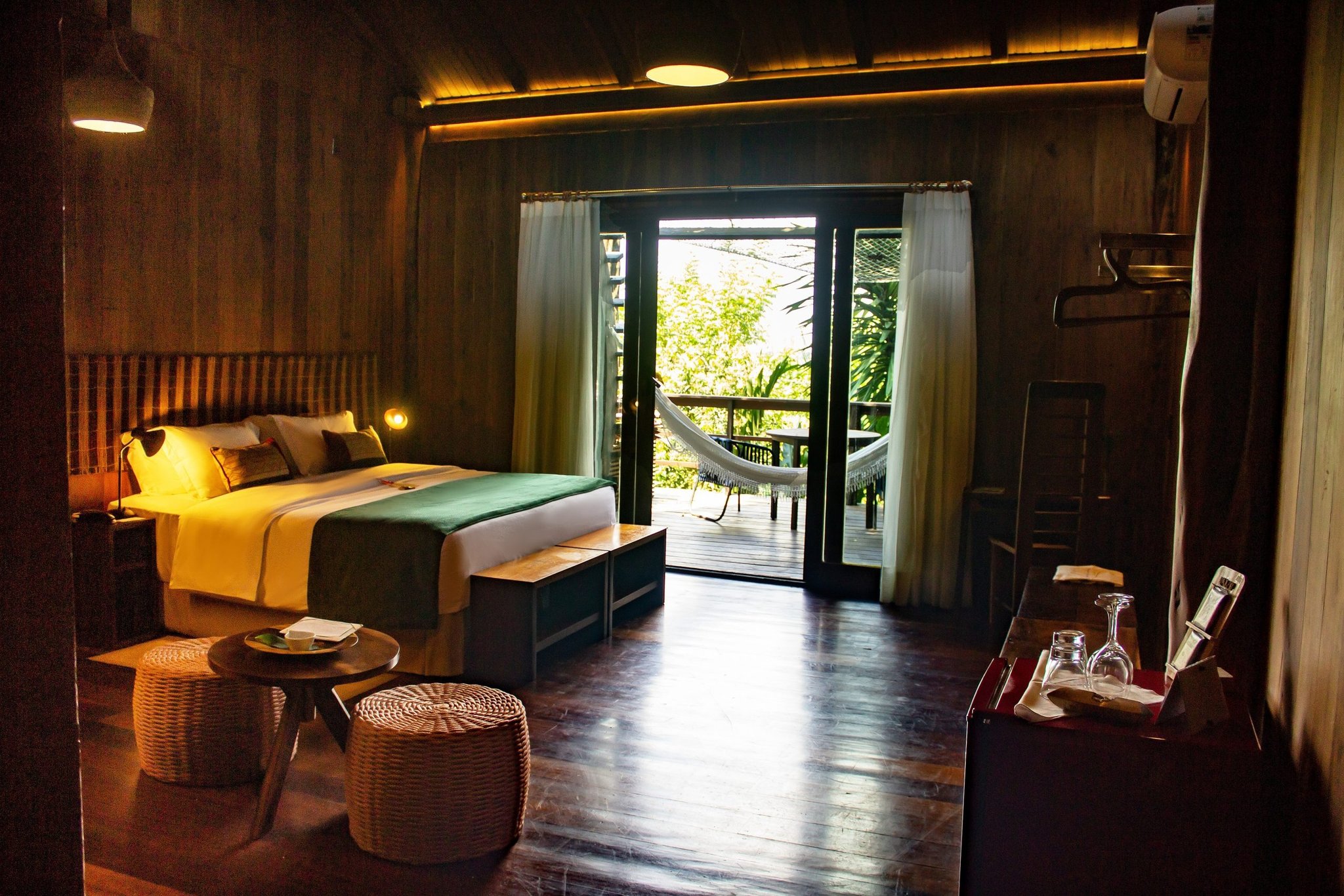 Your own hammock, a canopy view, and cool dark hardwood underfoot. Warmly lit luxury amazon lodge brazil suite with a double bed, wicker ottomans, and open French doors leading to a private hammock balcony surrounded by tropical greenery