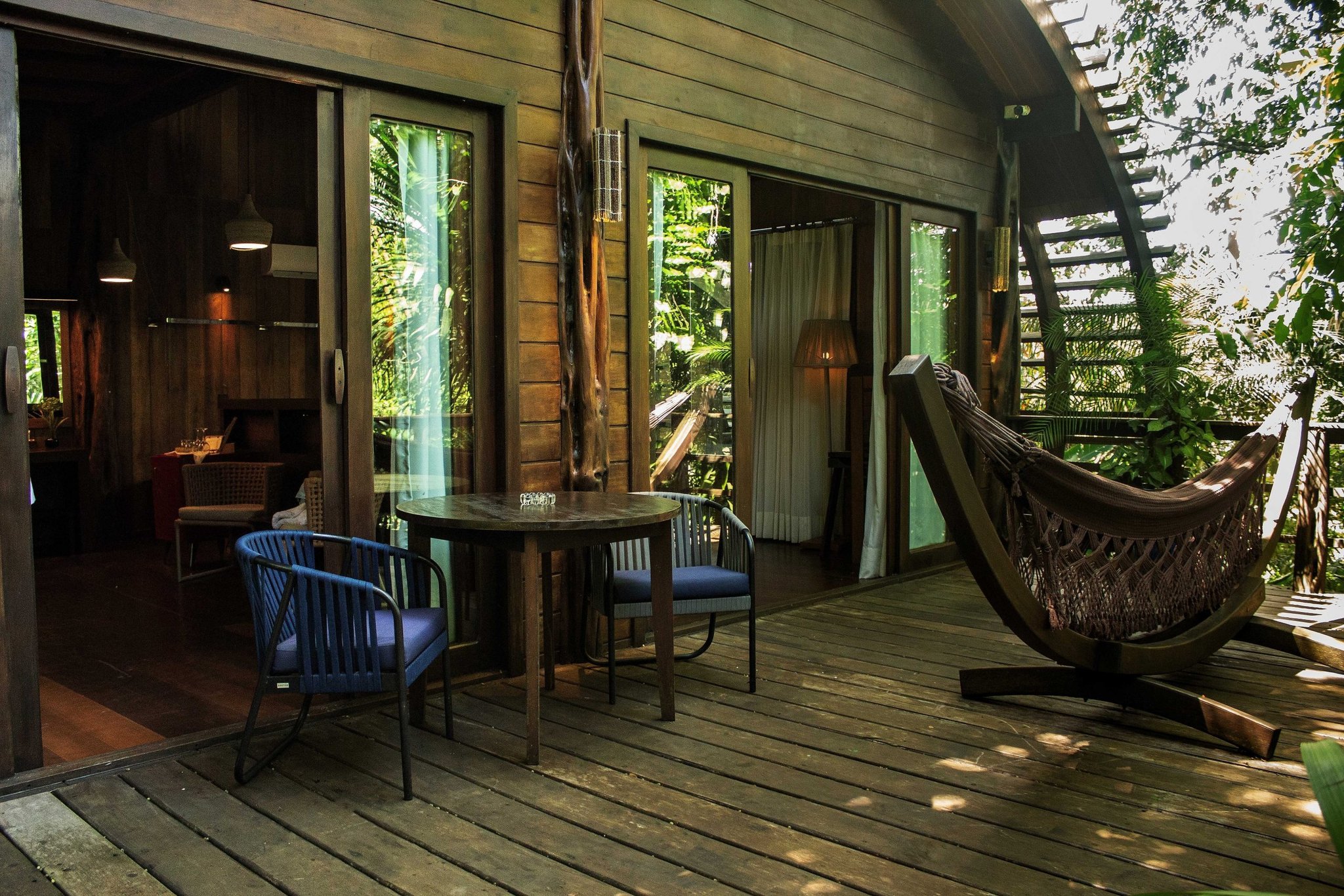Hammock, hardwood deck, jungle pressing in — slow down and stay a while. A private wooden deck at a luxury amazon lodge brazil featuring a round table with two chairs, a freestanding hammock, and sliding glass doors opening to a forest-surrounded cabin