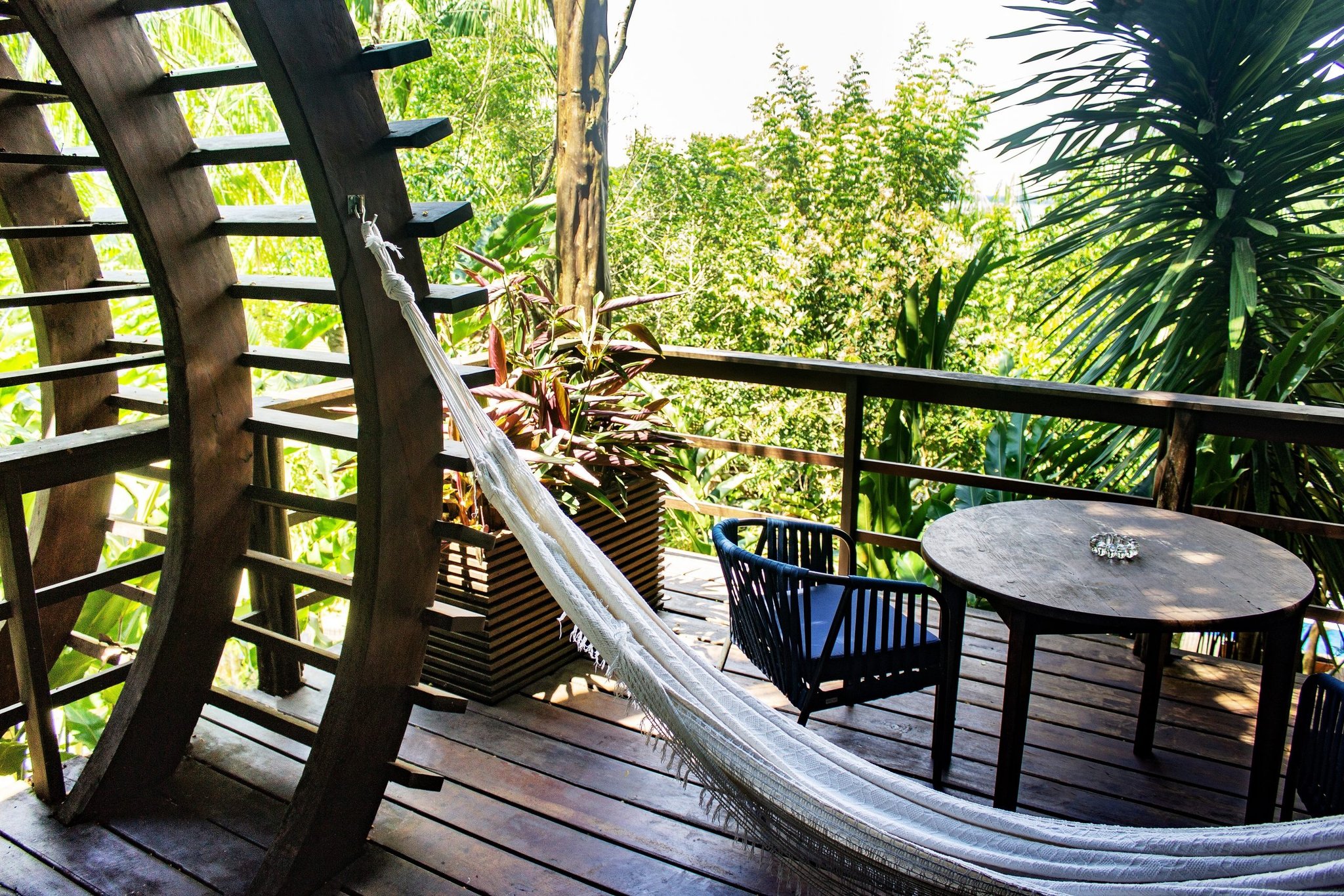 Afternoon light, a hammock, and the jungle pressing in from every side. A white fringed hammock and round hardwood table on a curved timber deck at a luxury amazon lodge brazil, surrounded by lush tropical greenery