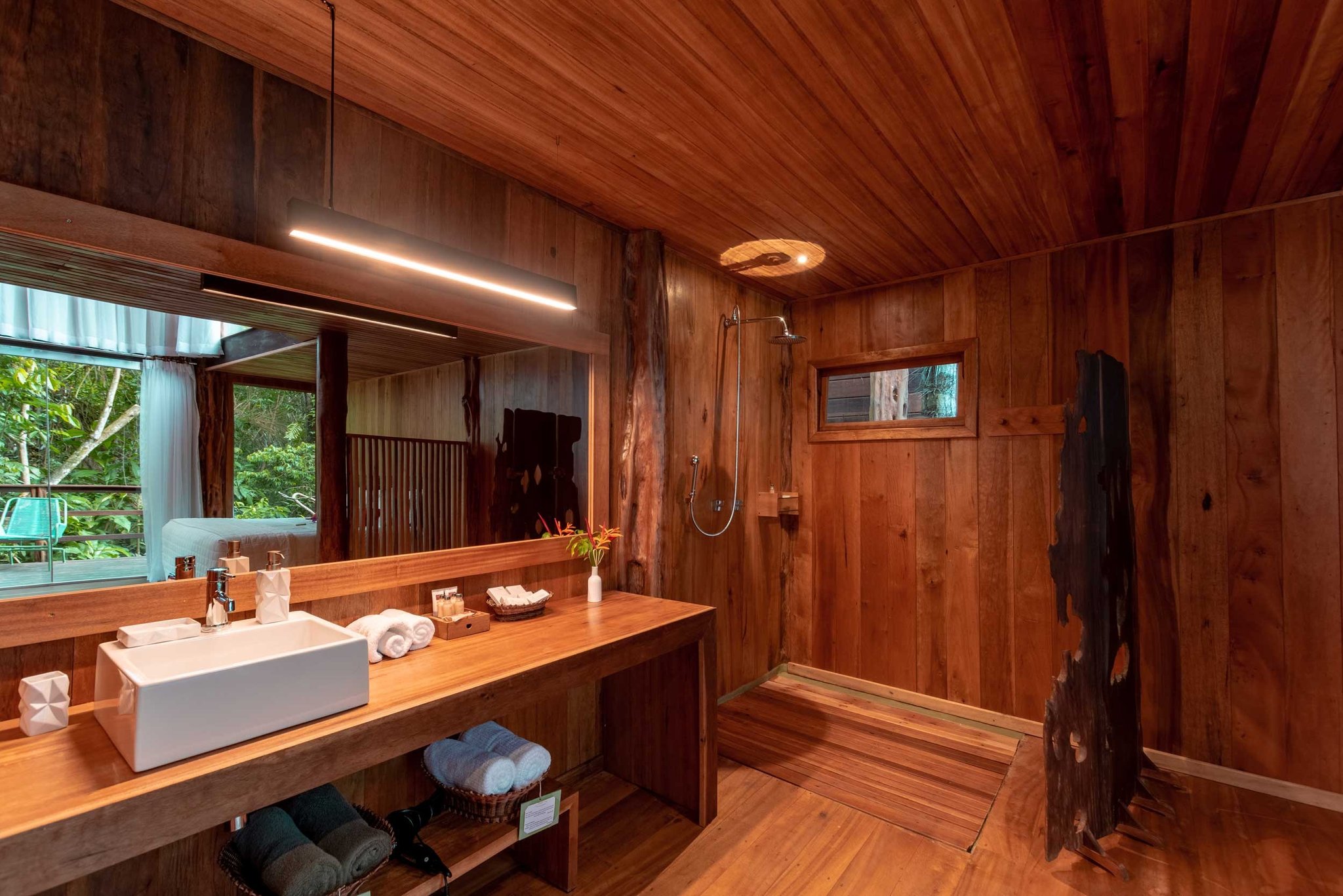 Rainforest greenery visible through the mirror, rustic timber wrapping every surface — comfort without compromise. Warmly lit ensuite bathroom at a luxury amazon lodge brazil, featuring floor-to-ceiling certified hardwood panelling, a vessel sink vanity, rainfall shower, and a large mirror reflecting lush jungle greenery outside