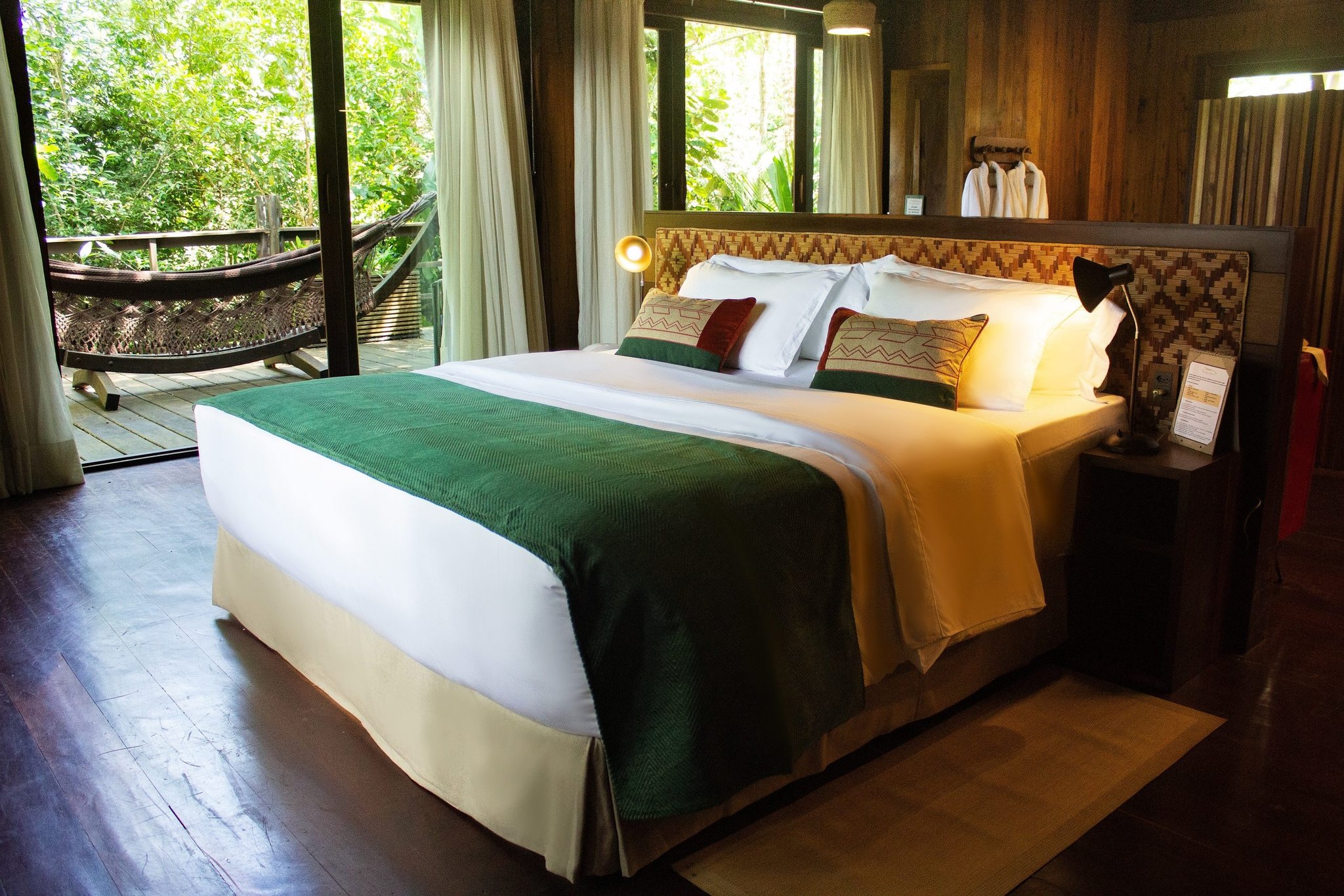 Crisp white linens, warm timber walls, a hammock waiting just beyond the glass. King bed with a woven geometric headboard and green velvet runner in a luxury amazon lodge brazil cabin, with a private hammock deck framed by lush rainforest visible through floor-to-ceiling glass doors