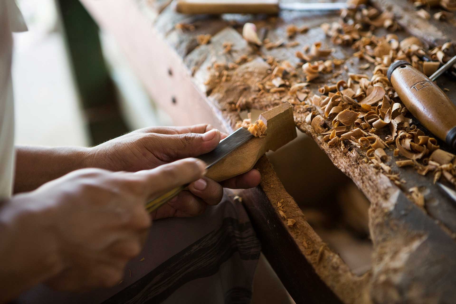 Curled shavings, steady hands — craftsmanship passed down through generations. Close-up of hands using a chisel to carve a wooden piece at a workbench covered in fresh wood shavings, reflecting the local artisan craft traditions near a luxury Amazon lodge in Brazil