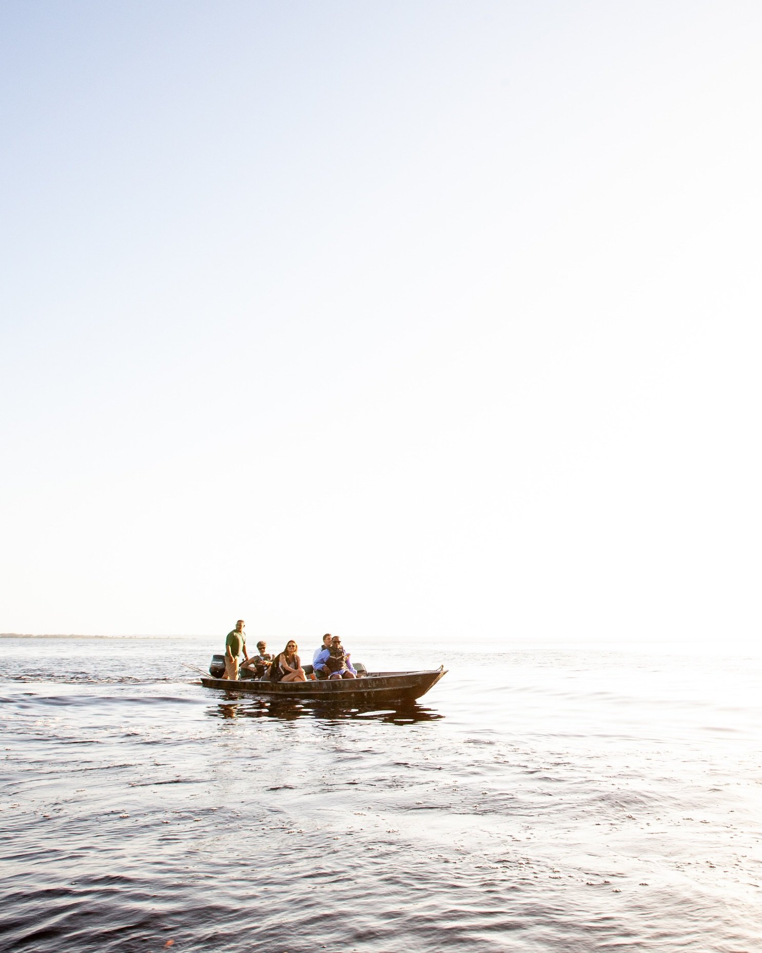 Five guests, one guide, endless river — the Amazon opens up around you. A small wooden motorboat carrying a group of travellers glides across a vast, calm expanse of dark river water under a bright hazy sky on a luxury Amazon lodge Brazil excursion
