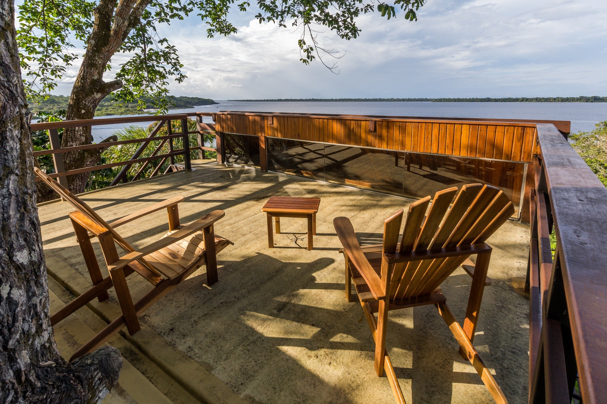 Settle in, pour a drink, let the river do the rest. Two wooden Adirondack chairs and a small side table on an open hardwood deck at a luxury Amazon lodge Brazil, overlooking a wide dark river bordered by dense rainforest under a cloudy sky