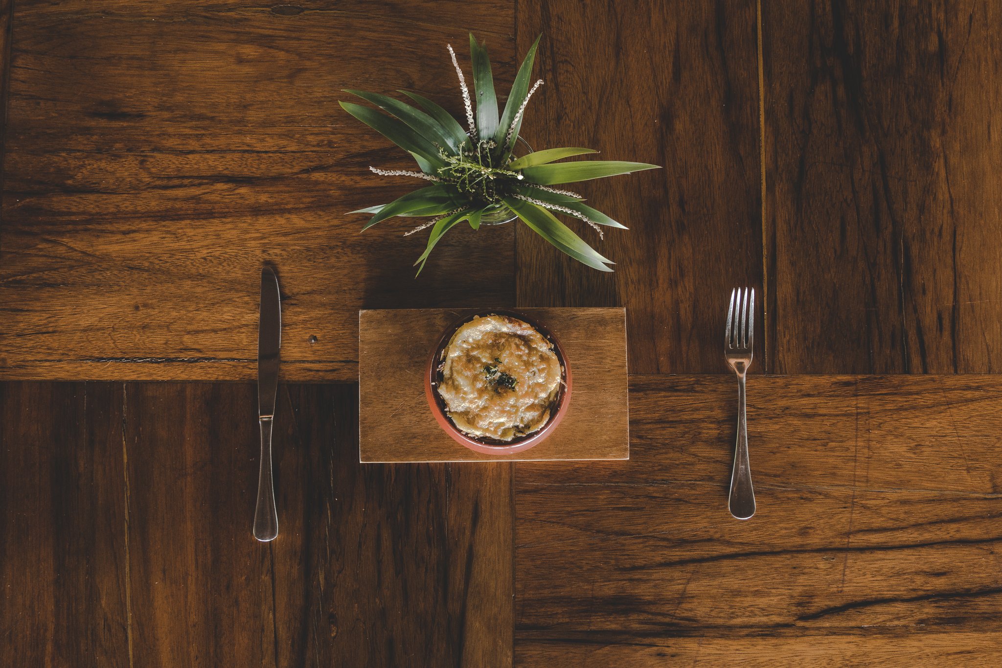 Simple, honest ingredients — plated with quiet elegance at the Camu-Camu Restaurant. Overhead flat-lay of a baked dish in a clay ramekin served on a wooden board with knife and fork, part of the luxury Amazon lodge Brazil dining experience