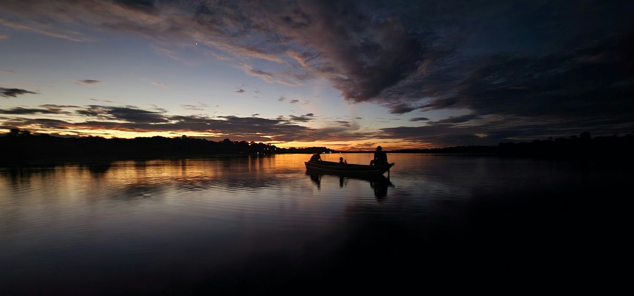 Three silhouettes, one boat, a sky on fire — dusk on the Rio Negro. Silhouette of three people in a small wooden boat on a glassy river at dusk, golden and violet clouds reflected on the still water near a luxury Amazon lodge Brazil experience