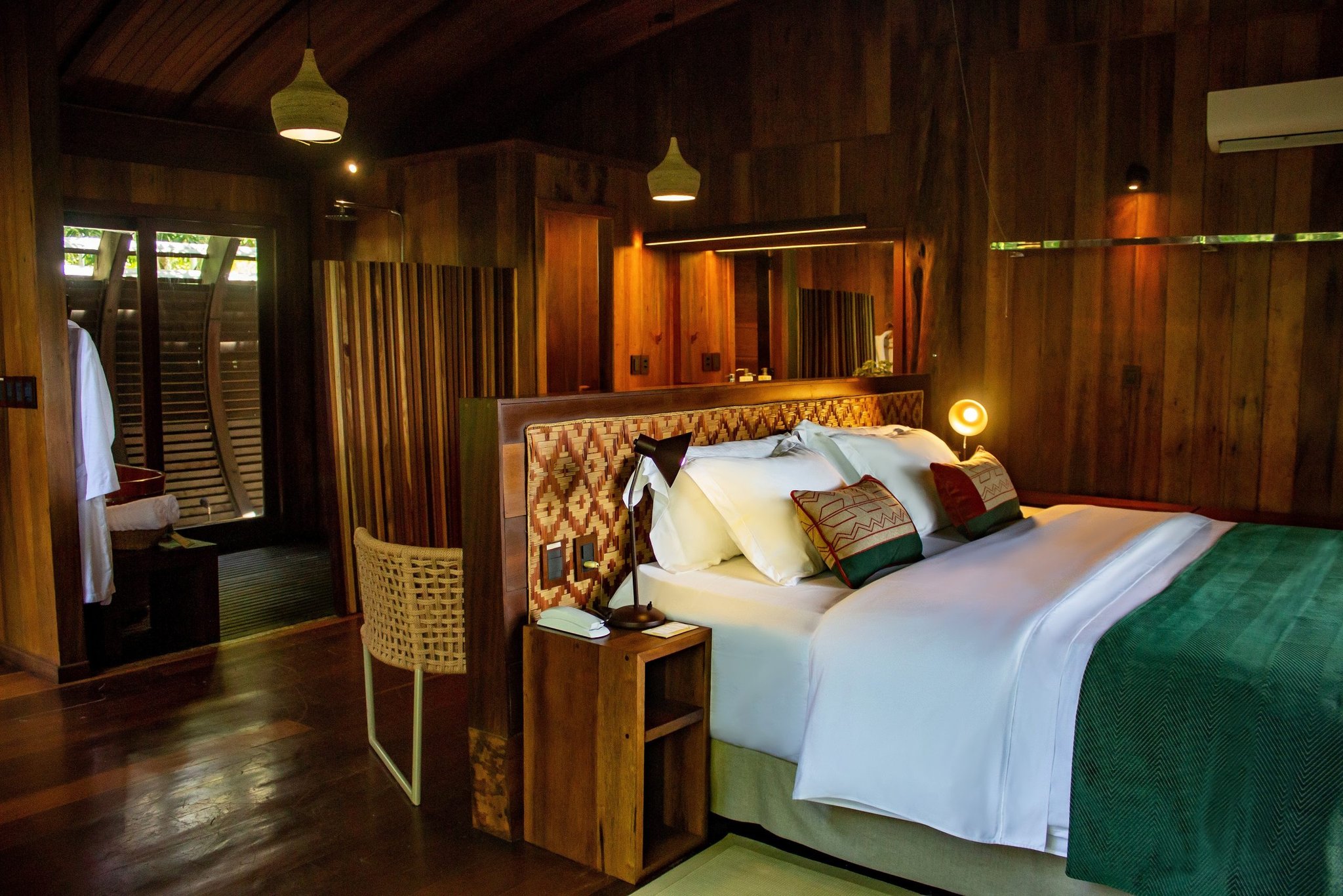 Crafted hardwood, handwoven details, crisp white linen — a suite that feels like the forest dreamed it. King bed with handwoven headboard and embroidered cushions inside a luxury Amazon lodge Brazil suite, surrounded by certified hardwood walls and warm pendant lighting