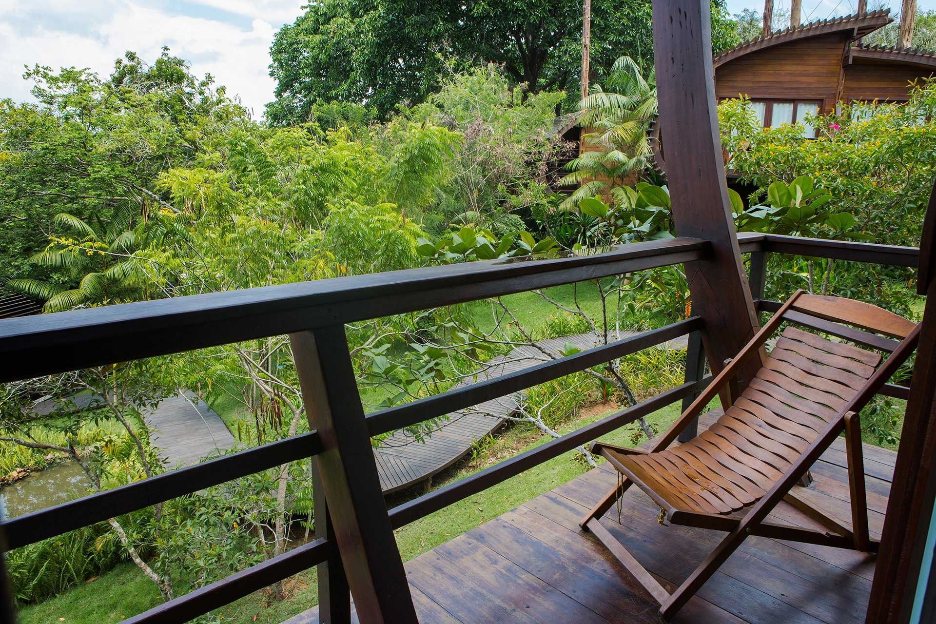 Your private perch above the green — jungle life unfolding just beyond the railing. A wooden deck chair sits on a private hardwood balcony at a luxury amazon lodge brazil, overlooking a lush canopy, timber walkways, and a neighbouring bungalow nestled among tropical trees