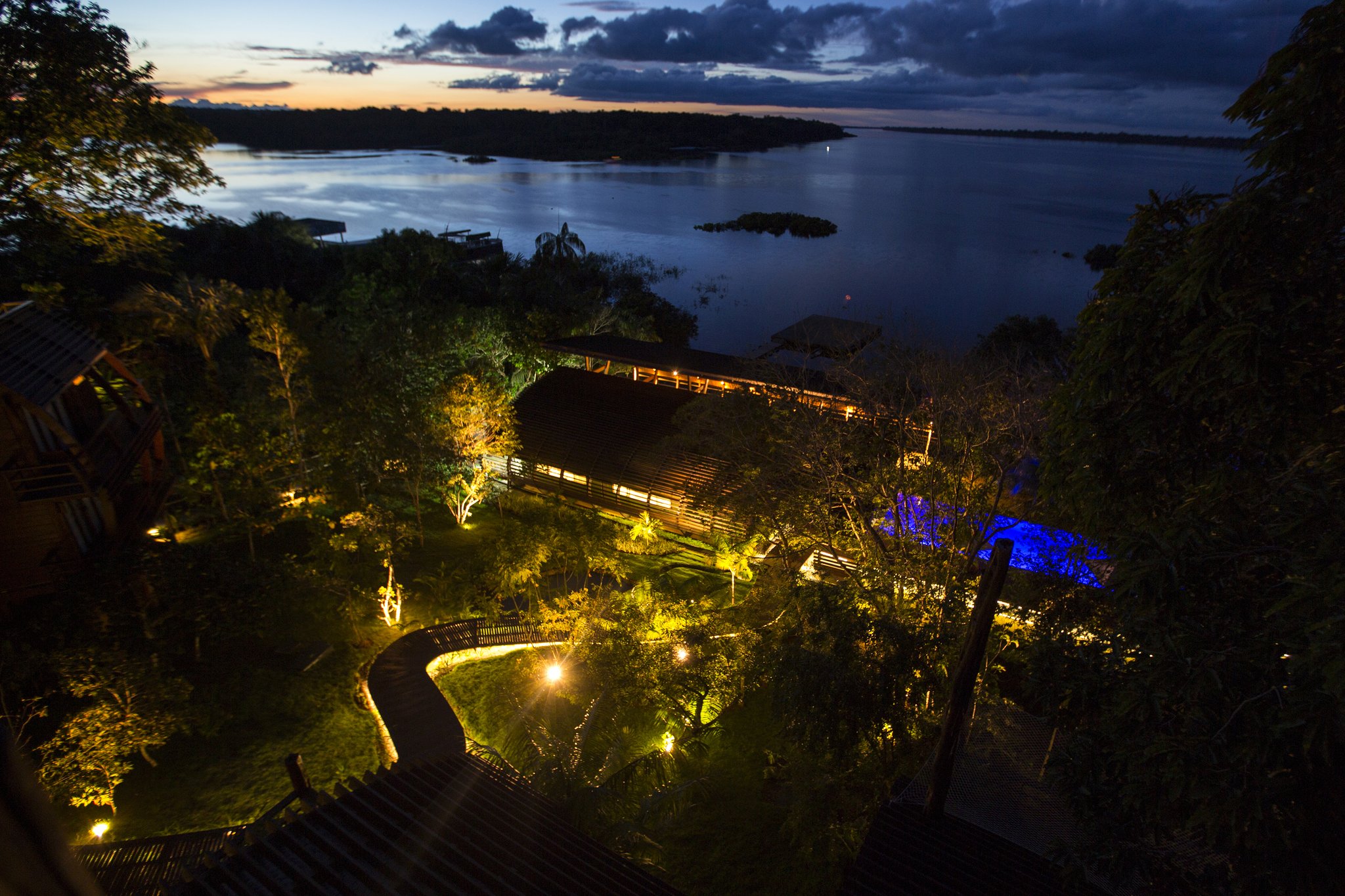 Jungle lights at dusk, a wide river stretching toward the horizon. Aerial twilight view of a luxury Amazon lodge Brazil, with warm golden lights illuminating timber buildings, a winding boardwalk and lush jungle garden leading down to a wide dark river dotted with forested islands under a dramatic dusk sky