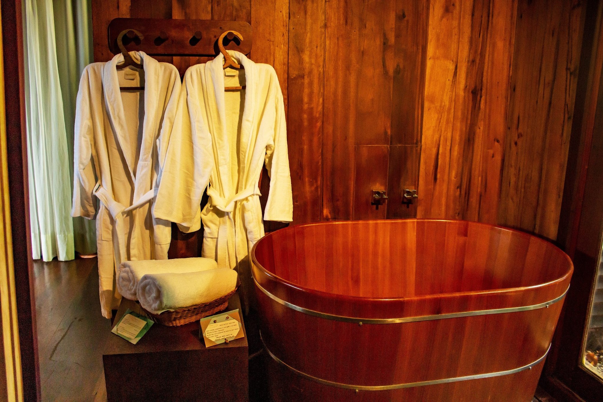 Soft robes, rolled towels, a handcrafted cedar tub — slow travel, Amazon-style. A polished round wooden soaking tub with brass fittings beside two white bathrobes hanging on a timber wall at a luxury Amazon lodge in Brazil