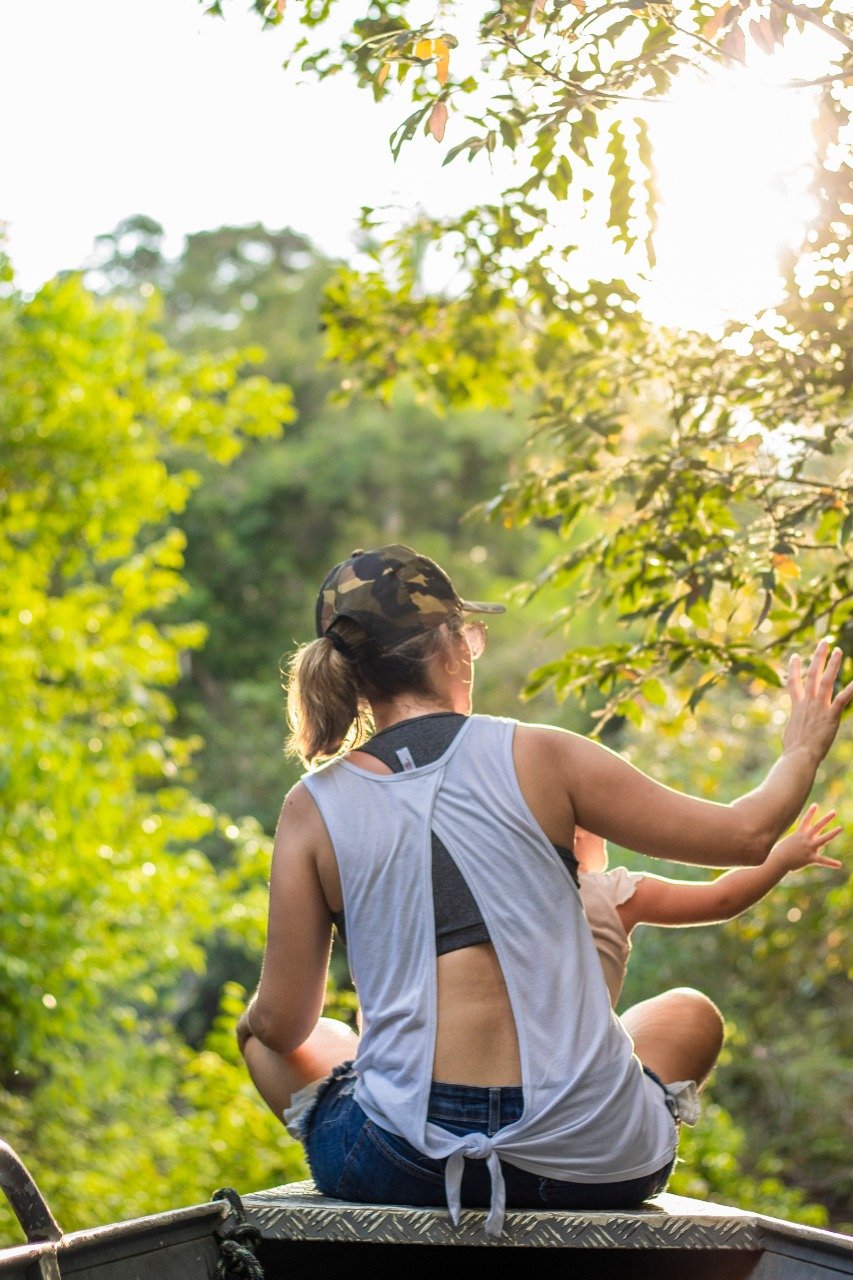 Sunlight, outstretched arms, and the forest pressing in from every side. A traveler sitting on the roof of a boat reaching toward overhanging tree branches during a manaus jungle tour, sunlight filtering through the rainforest canopy