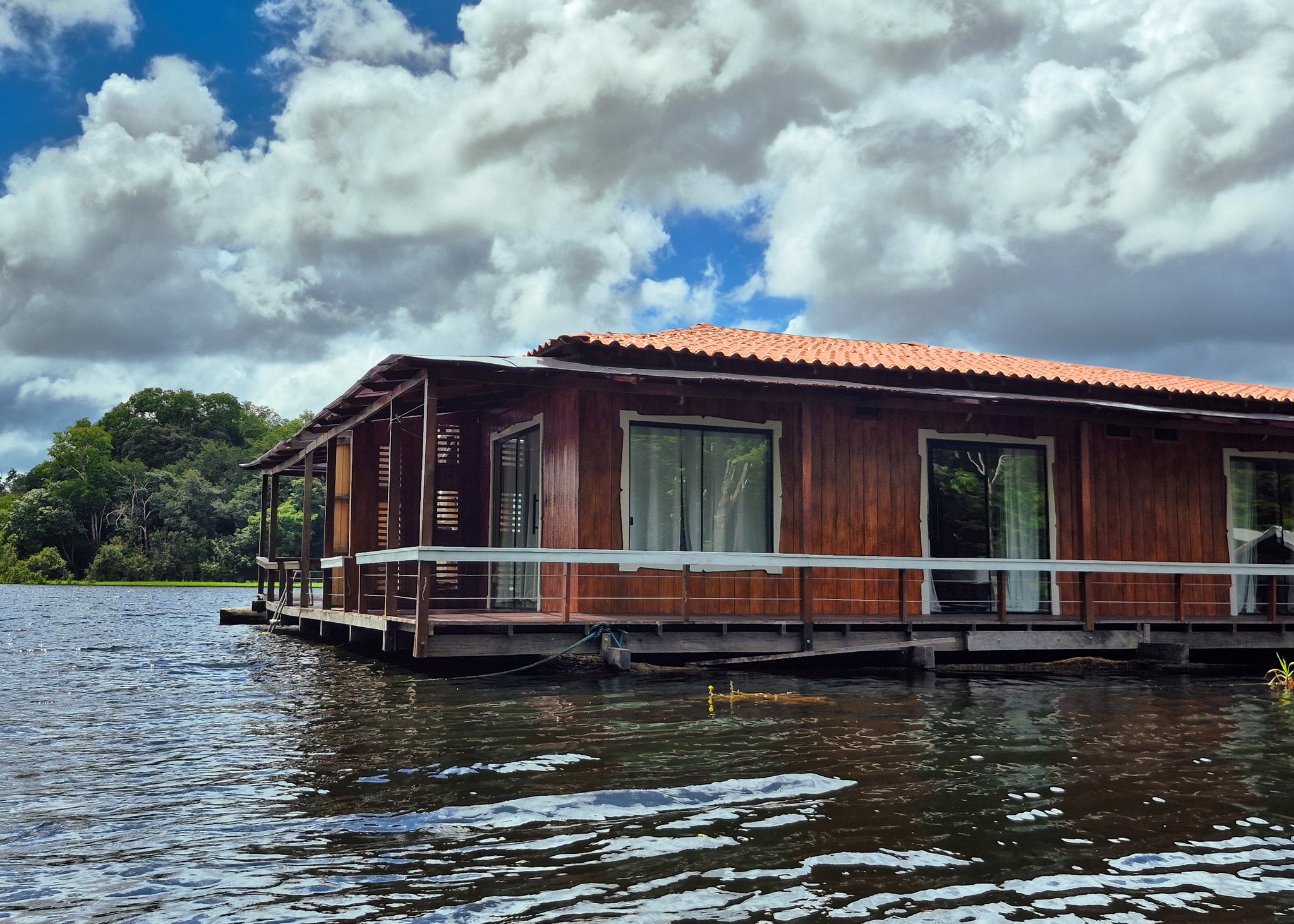 Sleeping on the water, deep in the rainforest. A wooden floating lodge with a terracotta-tiled roof and wraparound deck sits directly on a dark river, surrounded by dense rainforest canopy under a dramatic cloudy sky — a manaus jungle tour accommodation unlike any other