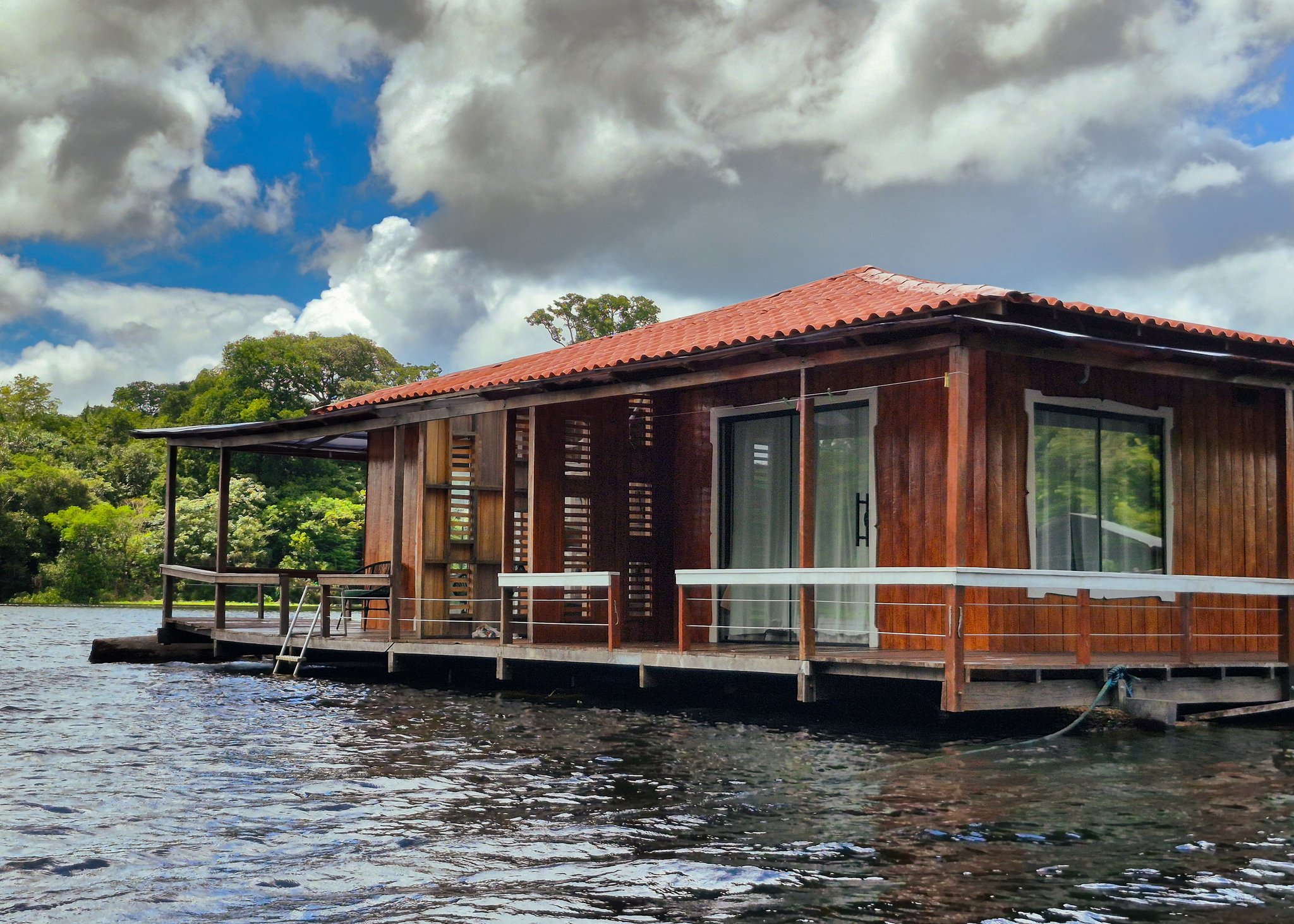 Your room for the night — a wooden cabin moored where the forest meets the river. A wooden floating lodge with a red-tiled roof and wraparound deck sits on dark Amazonian water, dense rainforest canopy visible on the shoreline behind it, on a manaus jungle tour