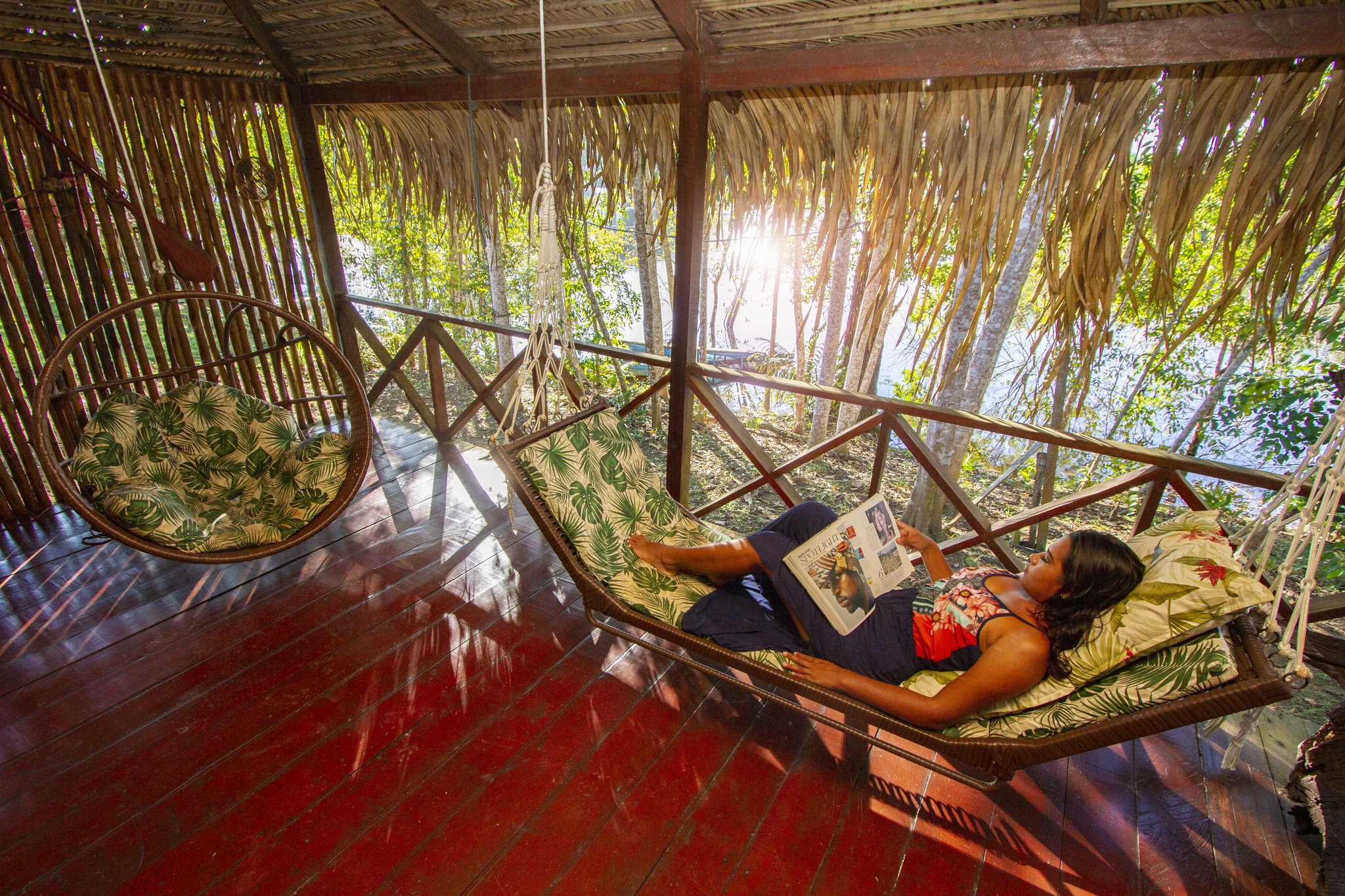 Pure jungle downtime — sunlight filtering through the palms while you swing and read. Photo: David Nascimento @ A woman lies in a suspended hammock swing reading a magazine on the open thatched-roof terrace of a jungle lodge, surrounded by tropical forest on a manaus jungle tour