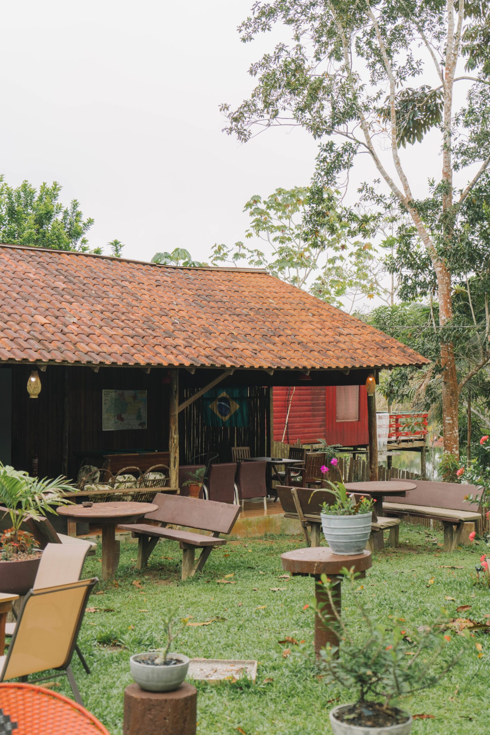 Wooden benches, potted flowers, and jungle trees all around — the lodge garden is yours to unwind in. Photo: Yuri Panichi Rustic open-air lodge common area with wooden benches and round tables on a green lawn, a terracotta-tiled roof and a Brazilian flag visible inside, surrounded by tropical trees on a manaus jungle tour