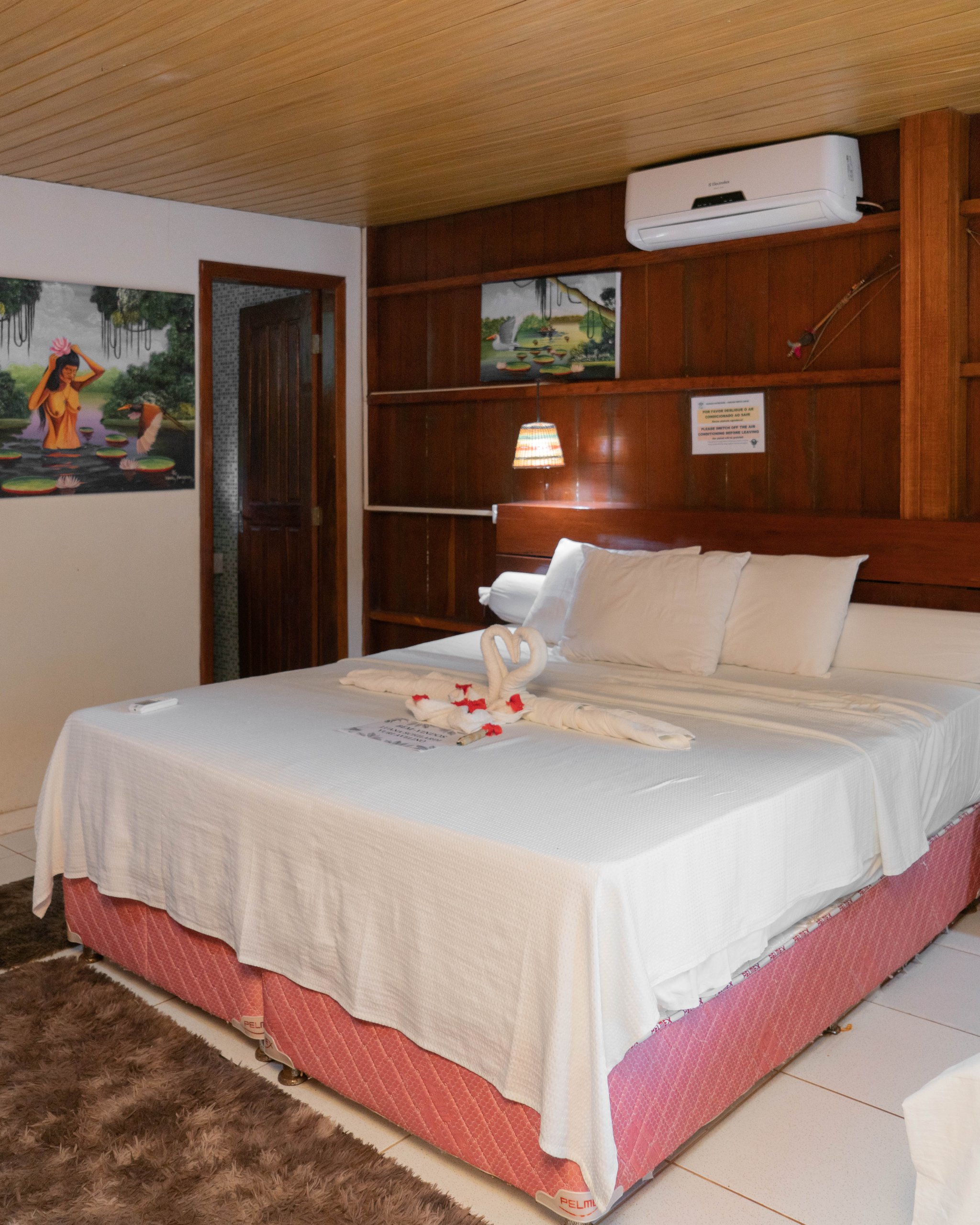 Towel swans and rose petals waiting after a full day in the rainforest. Photo: Yuri Panichi A neatly made double bed with towel swans and red flower petals at a jungle lodge on a manaus jungle tour, with wooden panelling, local artwork, and air conditioning