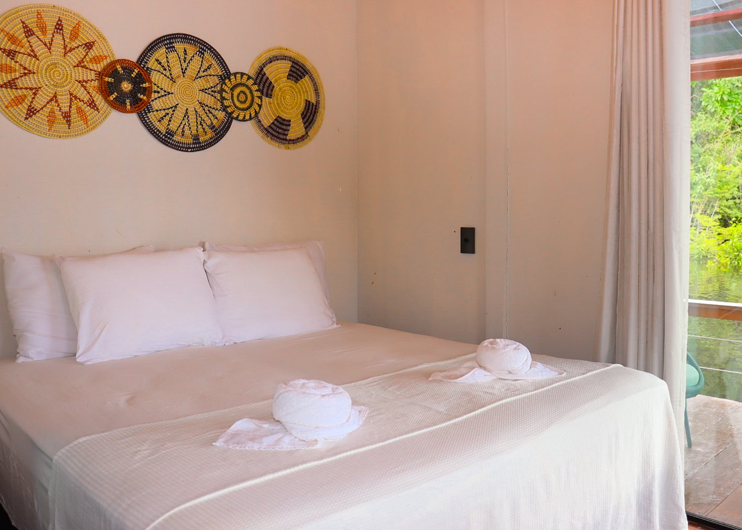 Handcrafted basket art, crisp white linens, and the green canopy just beyond the window. Double bed with white linens and folded towels in a jungle lodge room decorated with colourful woven basket wall art and a window overlooking lush green rainforest vegetation on a manaus jungle tour
