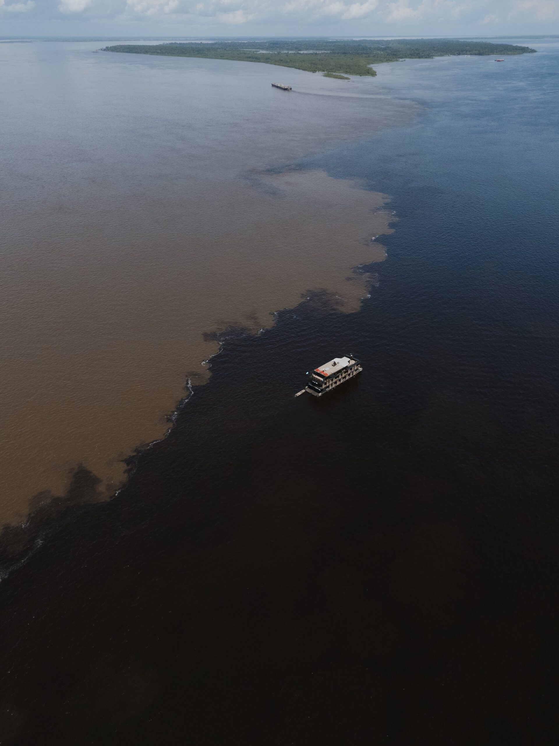 One boat, two rivers, zero mixing — nature's sharpest dividing line. Aerial view of a river vessel sailing the exact boundary line of the Meeting of the Waters on a manaus jungle tour, where dark black-water and sandy brown-water rivers flow side by side without mixing, with a forested island in the distance