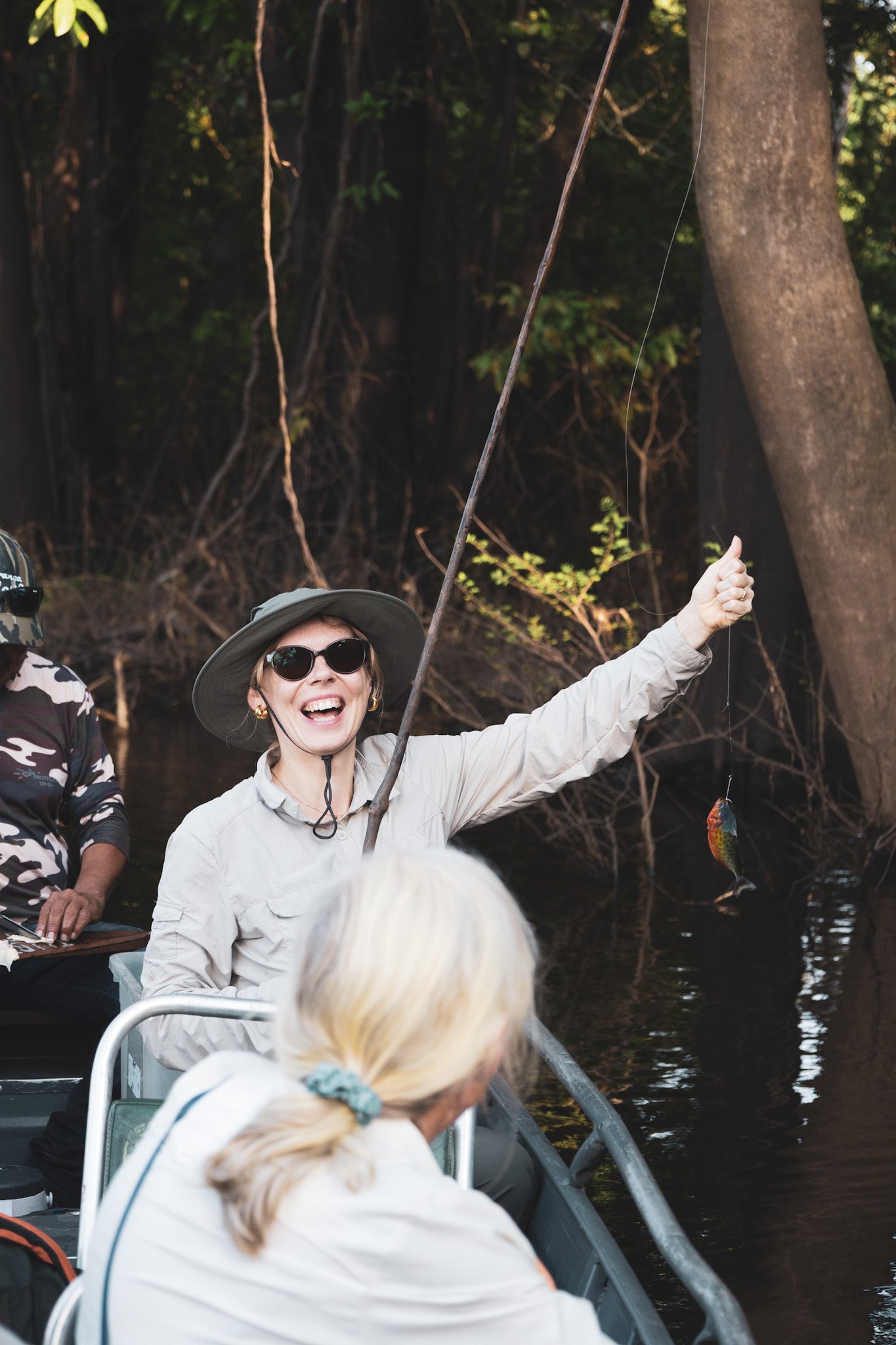 First piranha. Pure joy. A delighted woman in a sun hat holds up a small freshly caught piranha on a fishing line during a manaus jungle tour, seated in a motorized canoe on dark Amazonian waters surrounded by flooded forest