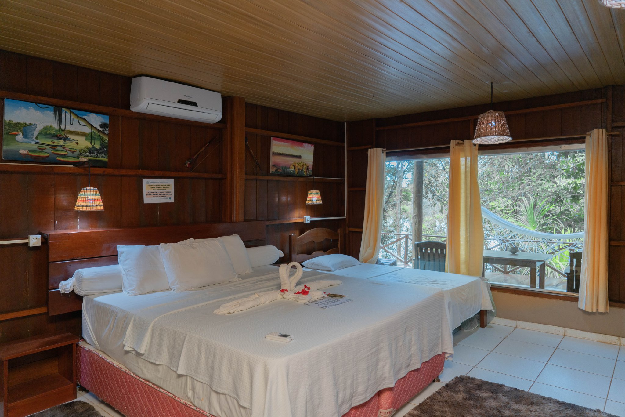 Cosy hardwood walls, a towel-swan welcome, and the Amazon forest just beyond the balcony. Photo: Yuri Panichi Wood-panelled twin bedroom on a manaus jungle tour, with white linens, towel-swan decoration, wicker pendant lamp, and a balcony overlooking dense forest canopy
