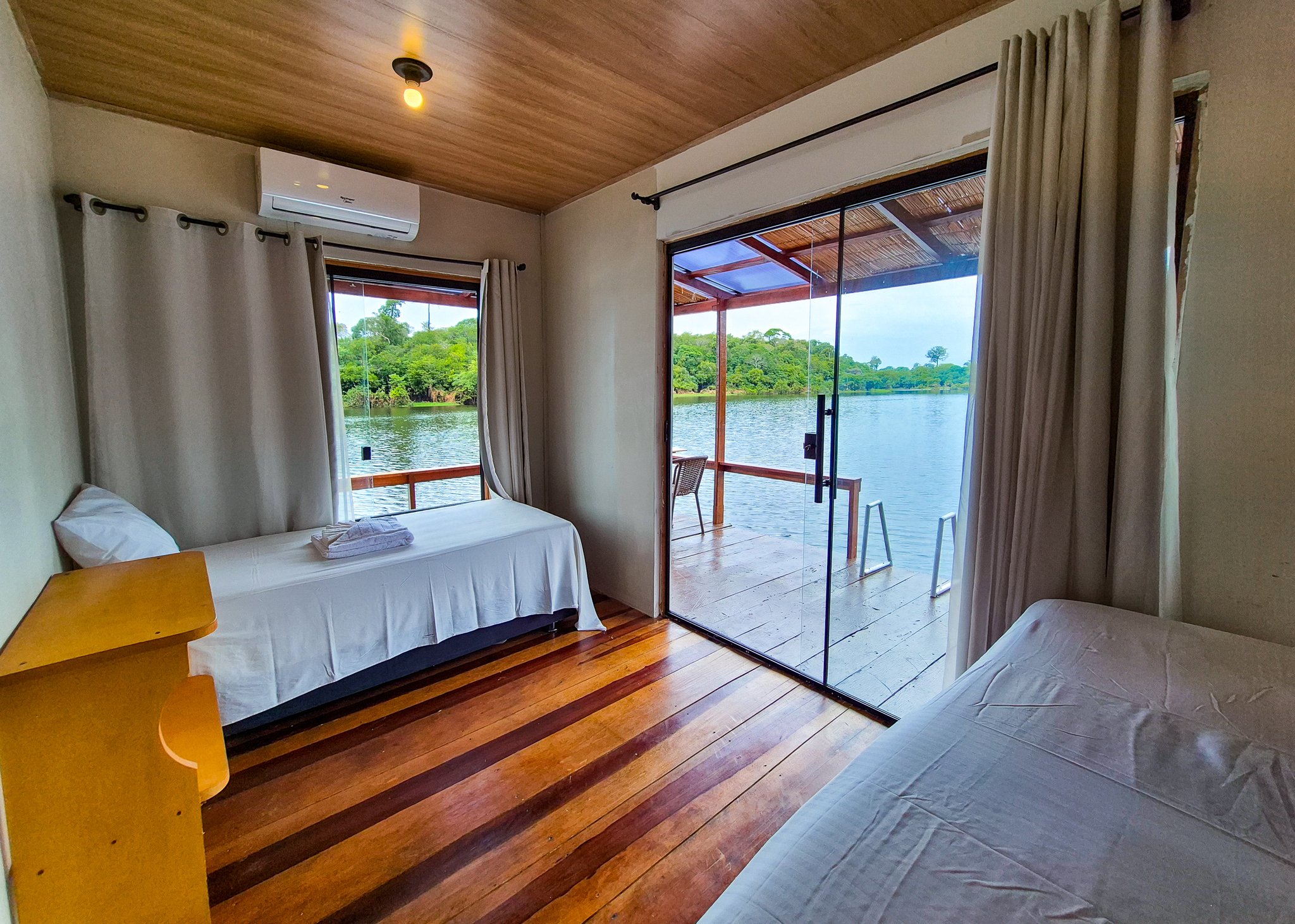 Fall asleep to the sound of the river, wake up to the forest. Twin-bed lodge room with polished hardwood floors and air conditioning, opening through a glass sliding door onto a covered deck with waterway and lush forest views — part of a manaus jungle tour accommodation