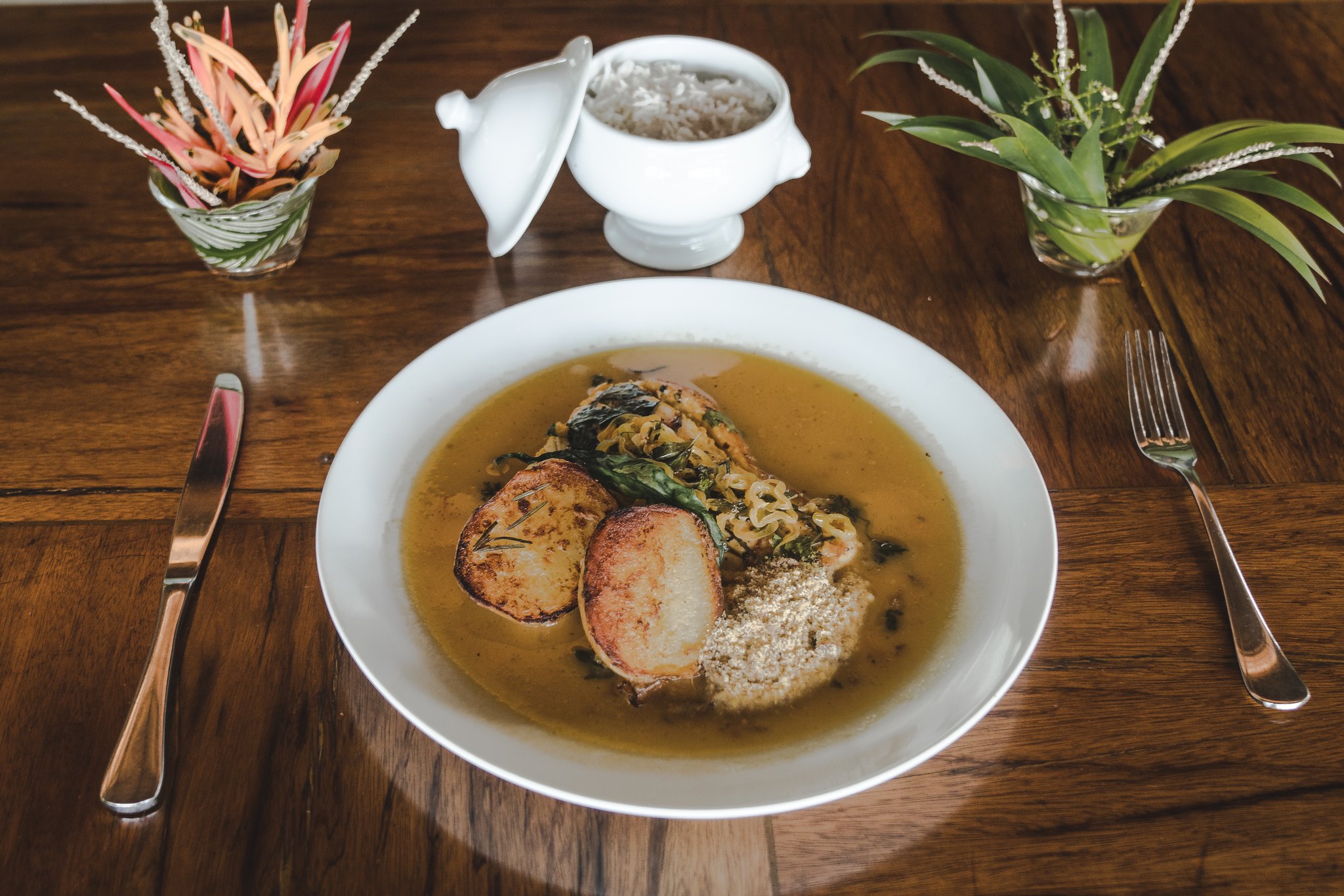 Farm-to-river flavours at the heart of the Amazon. A white bowl of Amazonian broth with seared medallions, greens, and cassava flour served at Mirante do Gavião lodge restaurant on a polished hardwood table, with a white tureen of rice and tropical floral arrangements