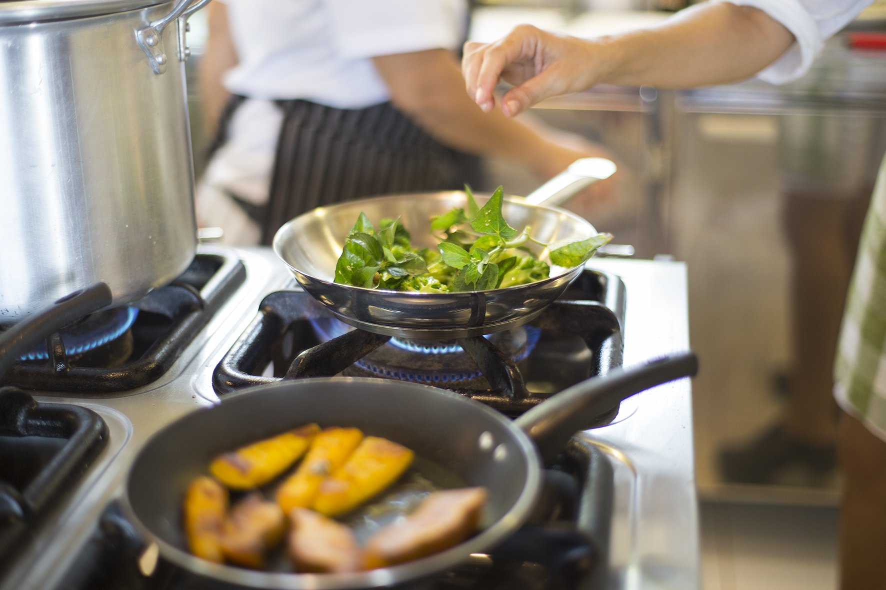 Farm-fresh flavours cooked to order at the lodge's Camu-Camu restaurant. A chef at Mirante do Gavião sautéing fresh green leaves in a steel pan over a gas flame, with caramelised fruit cooking in a second pan in the foreground