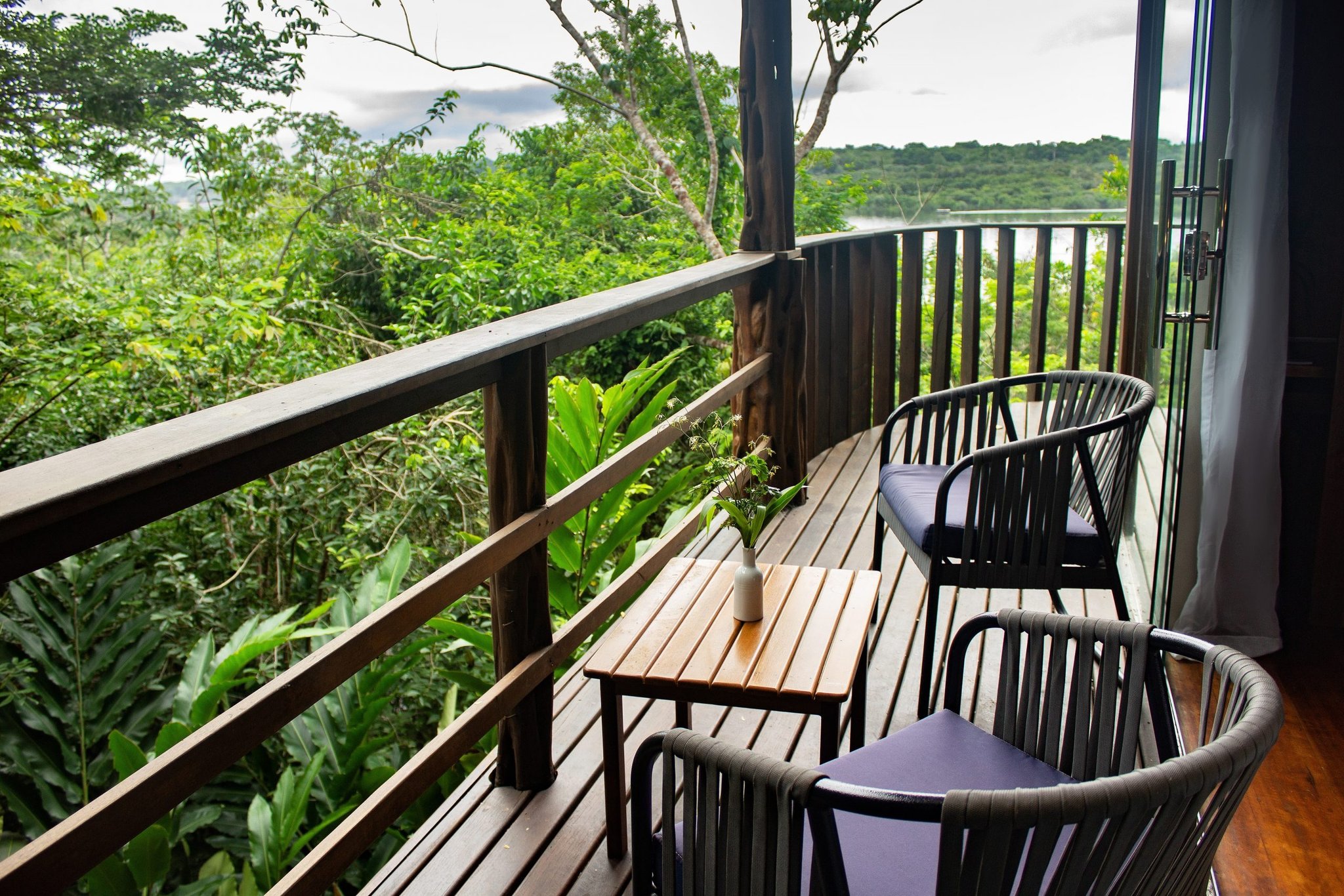Your own slice of the canopy — morning coffee never looked this good. Private curved wooden balcony at Mirante do Gavião with two cushioned chairs, a small teak table, and an unobstructed view over the Amazon rainforest canopy and Rio Negro