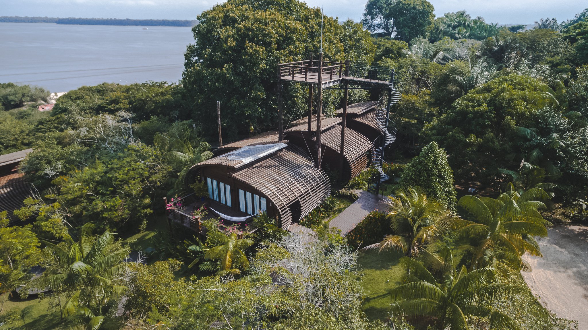 Where the jungle meets design — Mirante do Gavião from above. Aerial view of Mirante do Gavião lodge showing its curved wooden architecture nestled among dense Amazon rainforest trees beside the Rio Negro, with a canopy walkway and observation tower rising above the treetops