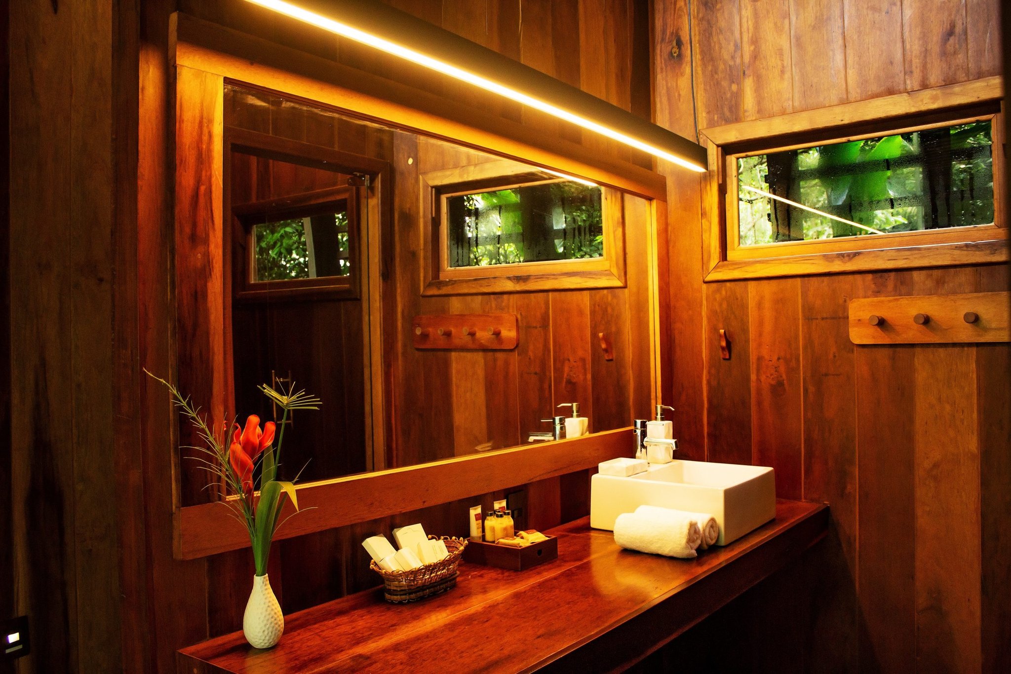 Jungle warmth meets refined comfort — every detail considered. Warm-lit private bathroom at Mirante do Gavião featuring a polished hardwood vanity, white vessel sink, folded towels, toiletry basket, and a tropical heliconia flower arrangement with jungle foliage visible through the windows