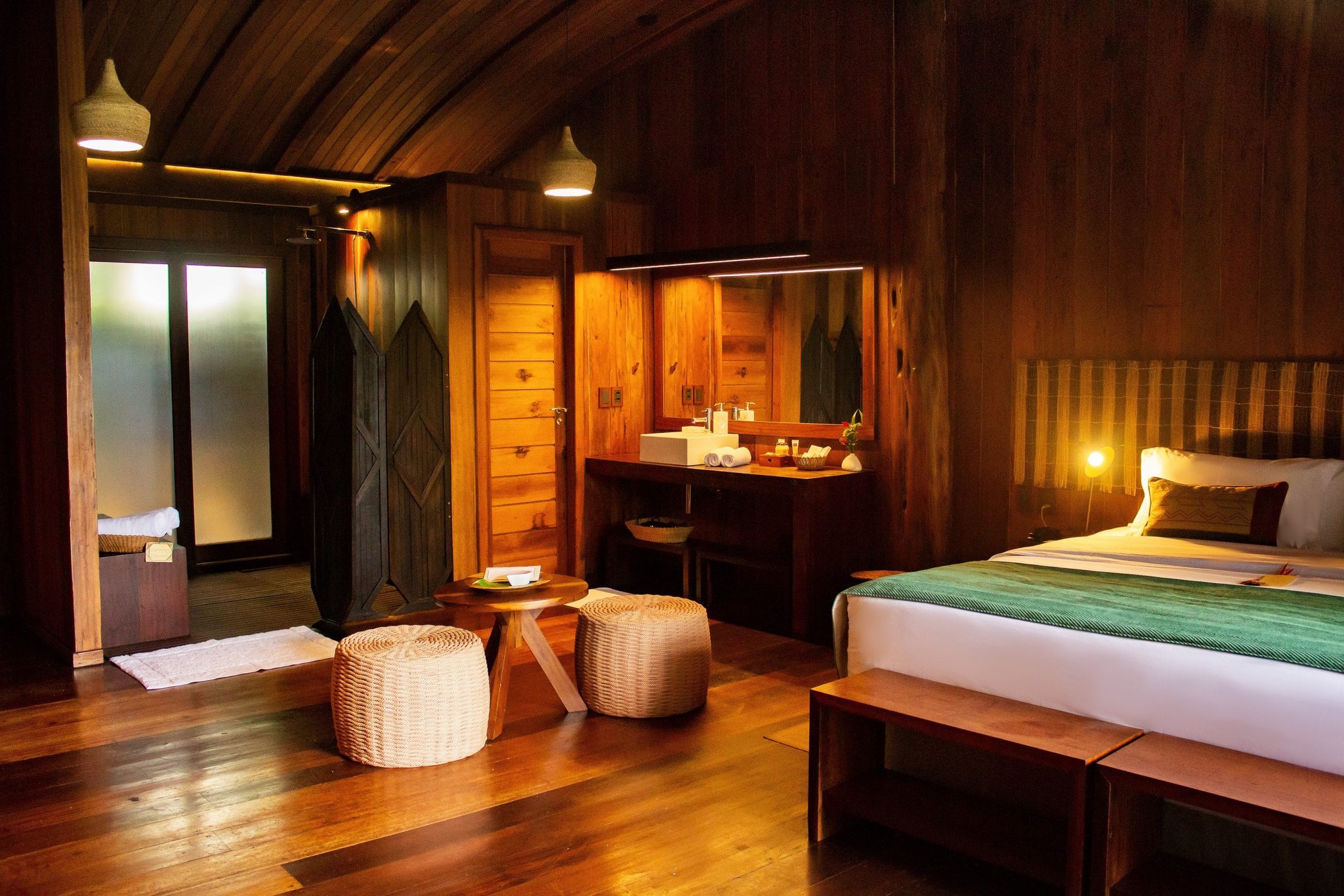 Rainforest comfort — every detail crafted from Amazonian hardwood. Warmly lit Mirante do Gavião lodge bedroom with polished hardwood floors, wicker stools, carved wooden wardrobe, vessel sink vanity, and a queen bed dressed in green linens