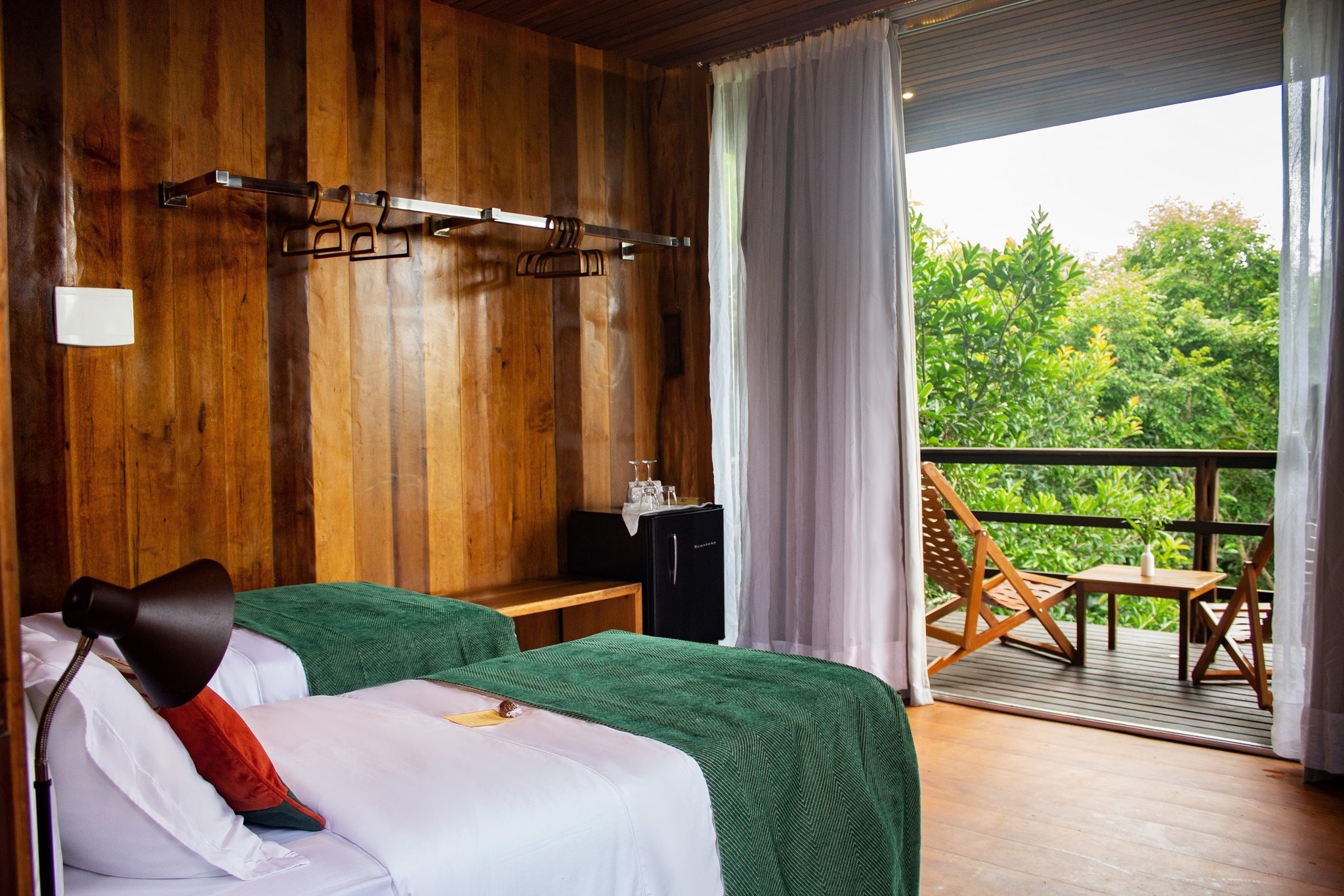 Wake up to the jungle — your private balcony at Mirante do Gavião. Mirante do Gavião lodge guest room with twin beds dressed in white linen and green throws, warm wooden plank walls, open glass door leading to a private balcony with teak loungers overlooking lush Amazon rainforest canopy