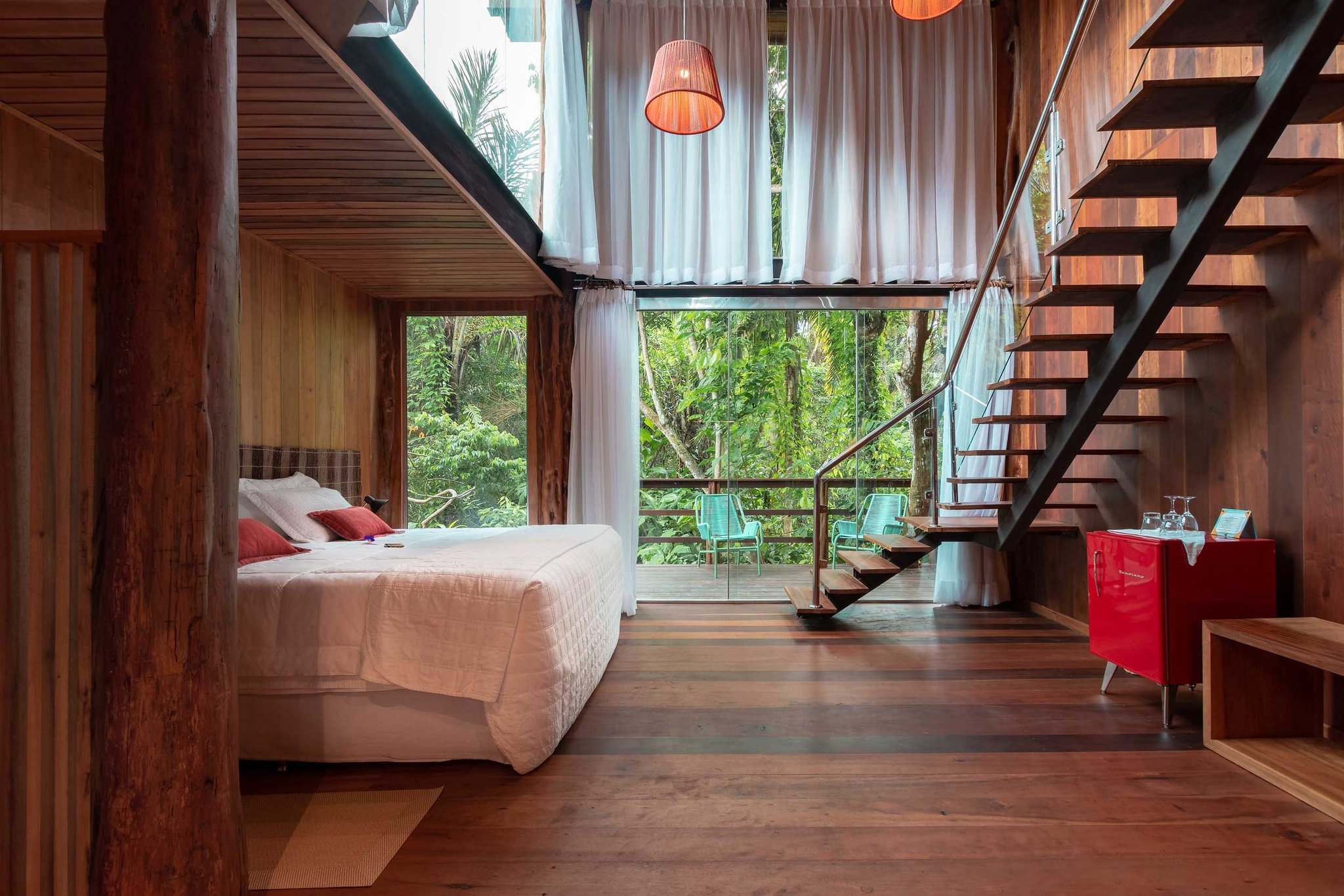 Wake up surrounded by Amazon jungle — your private balcony is just steps away. Warm timber-clad loft bedroom at Mirante do Gavião with a white double bed, floating staircase, red pendant lamp, and floor-to-ceiling glass doors opening onto a rainforest balcony